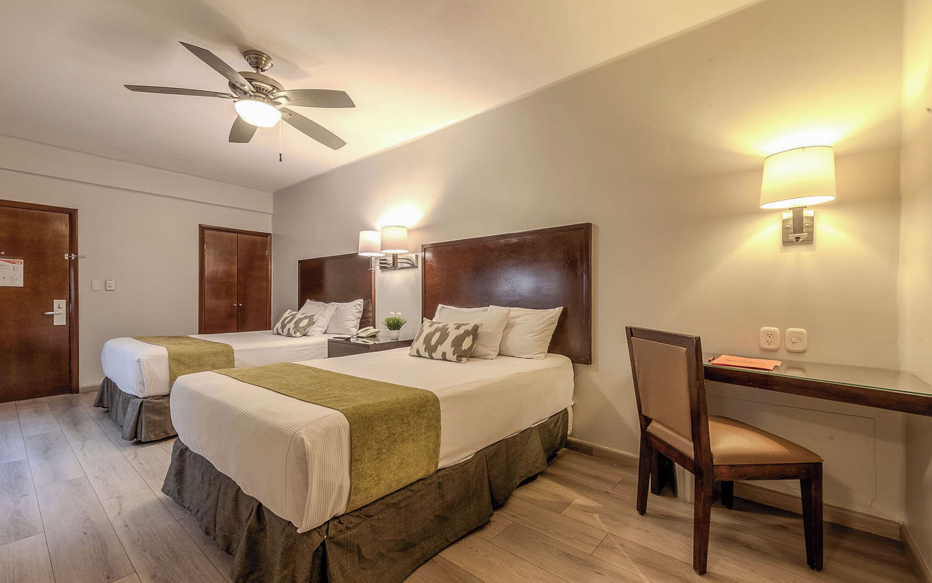 Ein Hotelzimmer mit zwei Betten, einem Schreibtisch, einem Stuhl, einem Deckenventilator und Wandlampen.