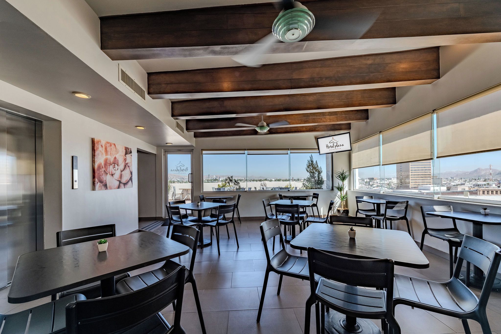Restaurante moderno com vista para a cidade, destacando vigas de madeira no teto, cadeiras pretas e mesas.