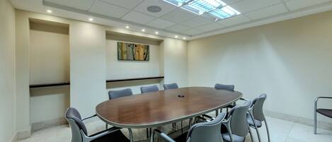 Sala de conferência com mesa redonda, seis cadeiras, iluminação embutida no teto e um quadro fixado na parede.