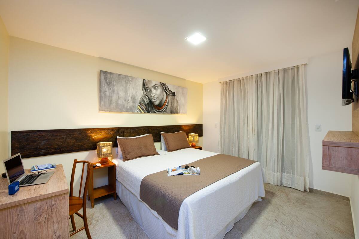 A hotel room with a bed, a desk with a laptop, a chair, a TV mounted on the wall, and a painting above the bed.