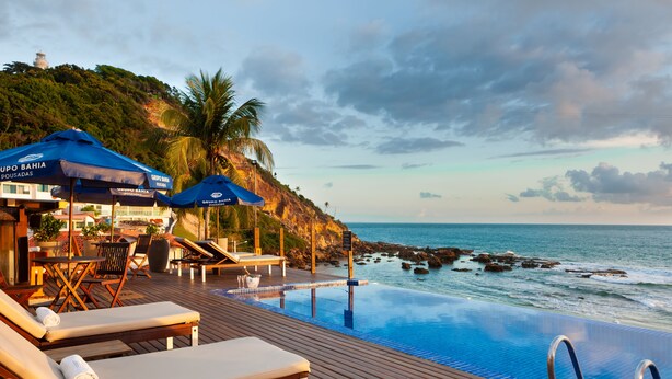 Una piscina infinita con sillones y sombrillas, con vista a un litoral rocoso y al océano.
