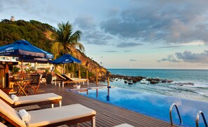 Una piscina infinita con sillones y sombrillas, con vista a un litoral rocoso y al océano.