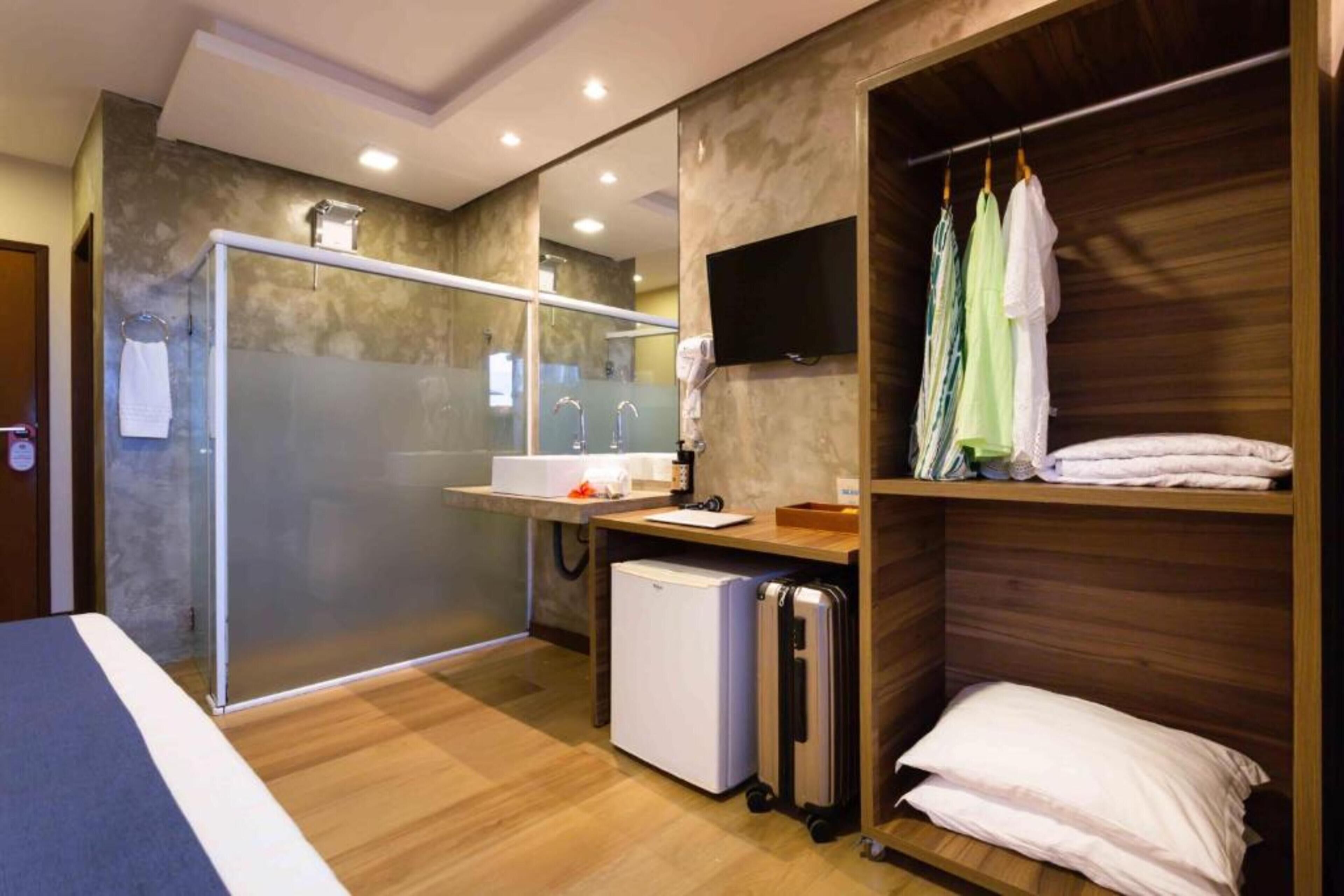 Une chambre d’hôtel équipée d’une douche, d’un petit réfrigérateur, d’une table de chevet, d’un téléviseur fixé au mur et d’une armoire avec des vêtements suspendus.