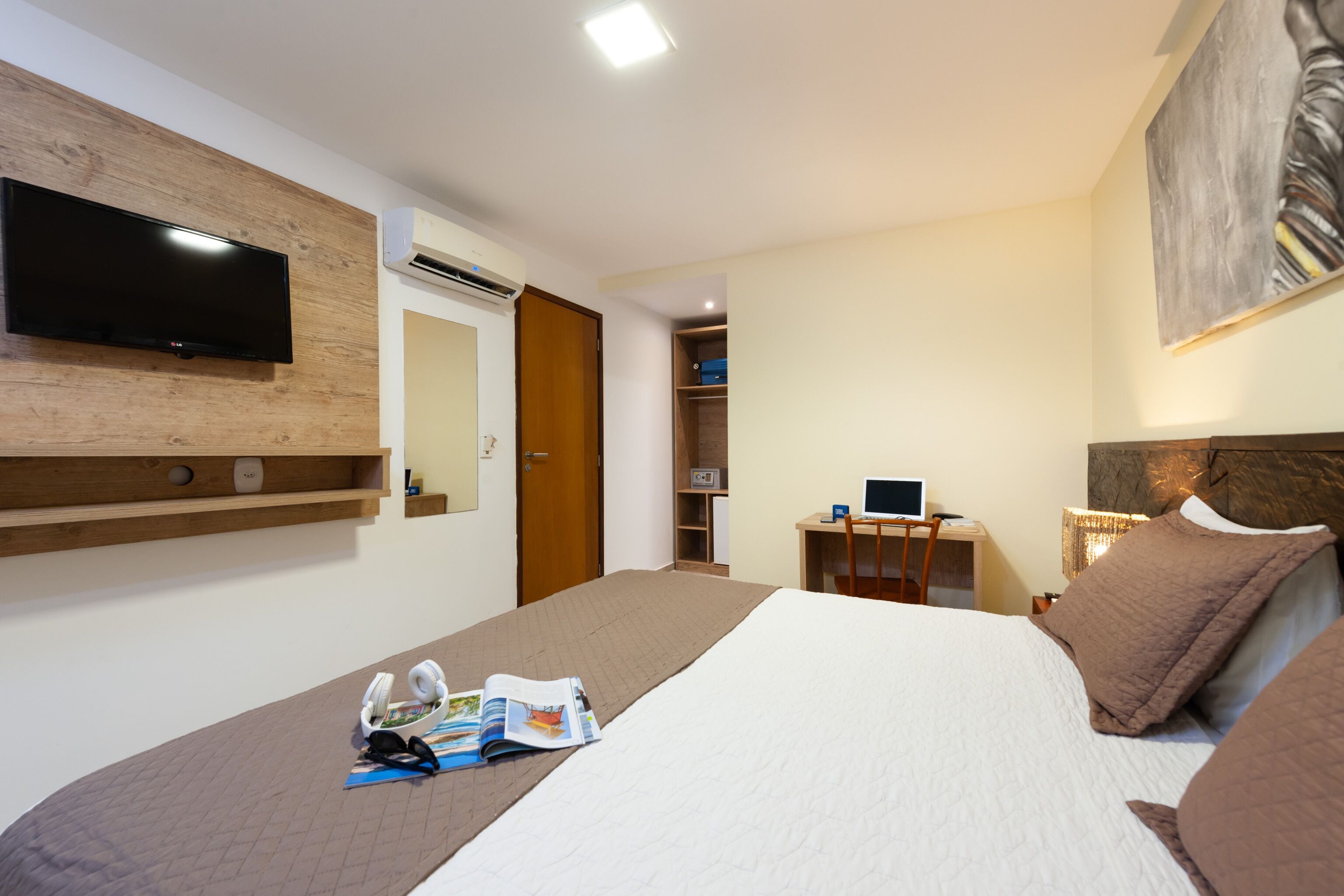 A hotel room with a bed, a desk, a chair, a flat-screen TV mounted on the wall, and a bookshelf.