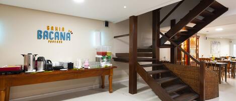 A modern interior with a wooden table displaying breakfast items, a staircase, and a wall with a sign reading "BAHIA BACANA."