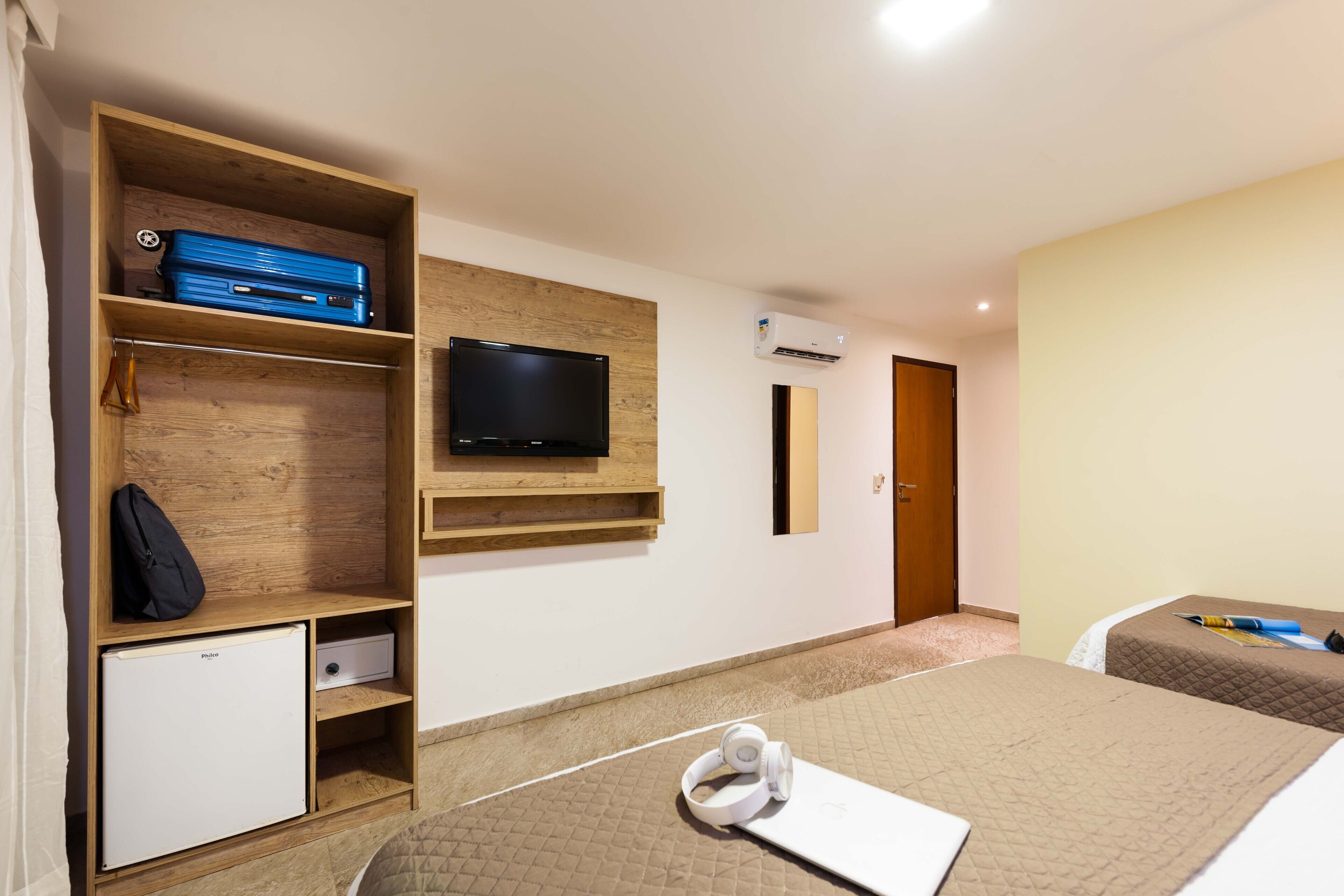 Hotelkamer met televisie, kleine koelkast, houten plank met een koffer, bed met quilt en nachtkastje met een boek en hoofdtelefoon.