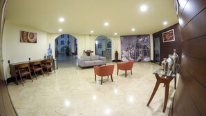 A spacious lobby with a marble floor, a sofa, two armchairs, a glass table, and a reception desk with chairs. - Hotel River Poliforum (León)