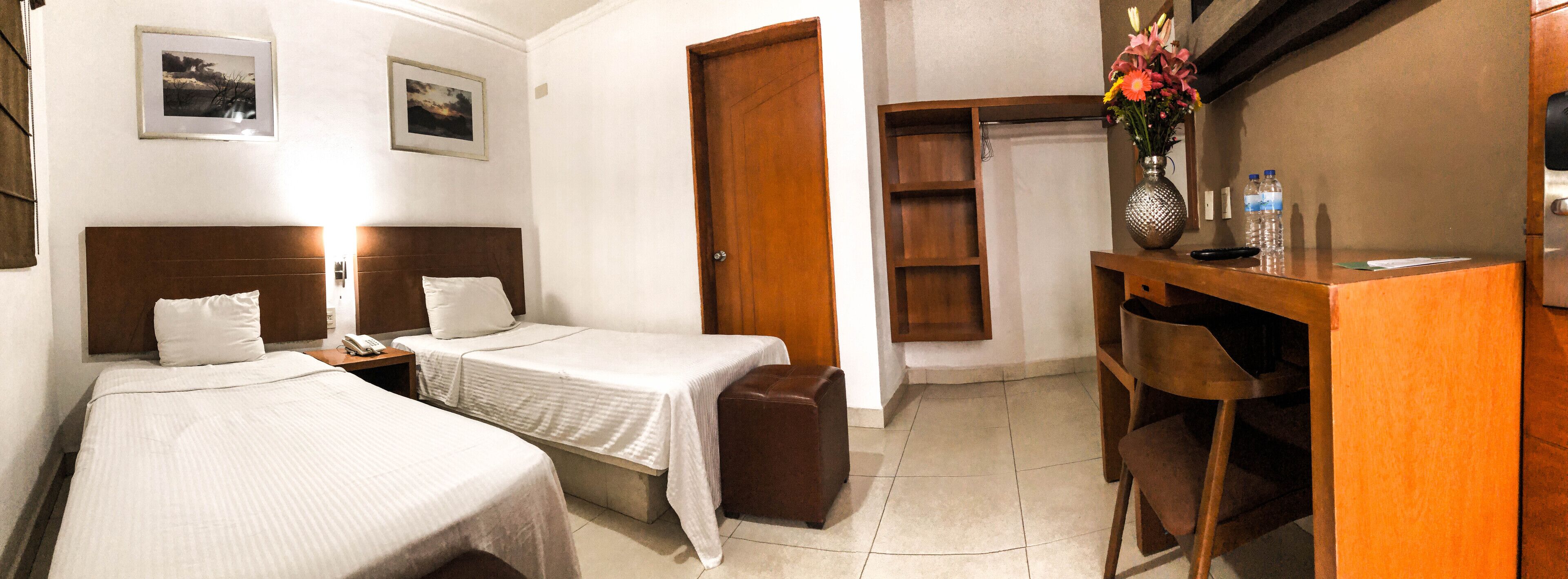 A hotel room with two single beds, a wooden desk with a vase of flowers, a chair, and a small cabinet.