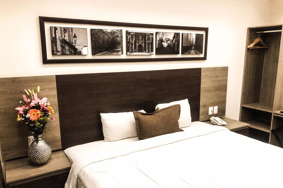 A hotel room with a large bed, a nightstand with a vase of flowers, a framed photo collage on the wall, and a wardrobe with a hanging rack.
