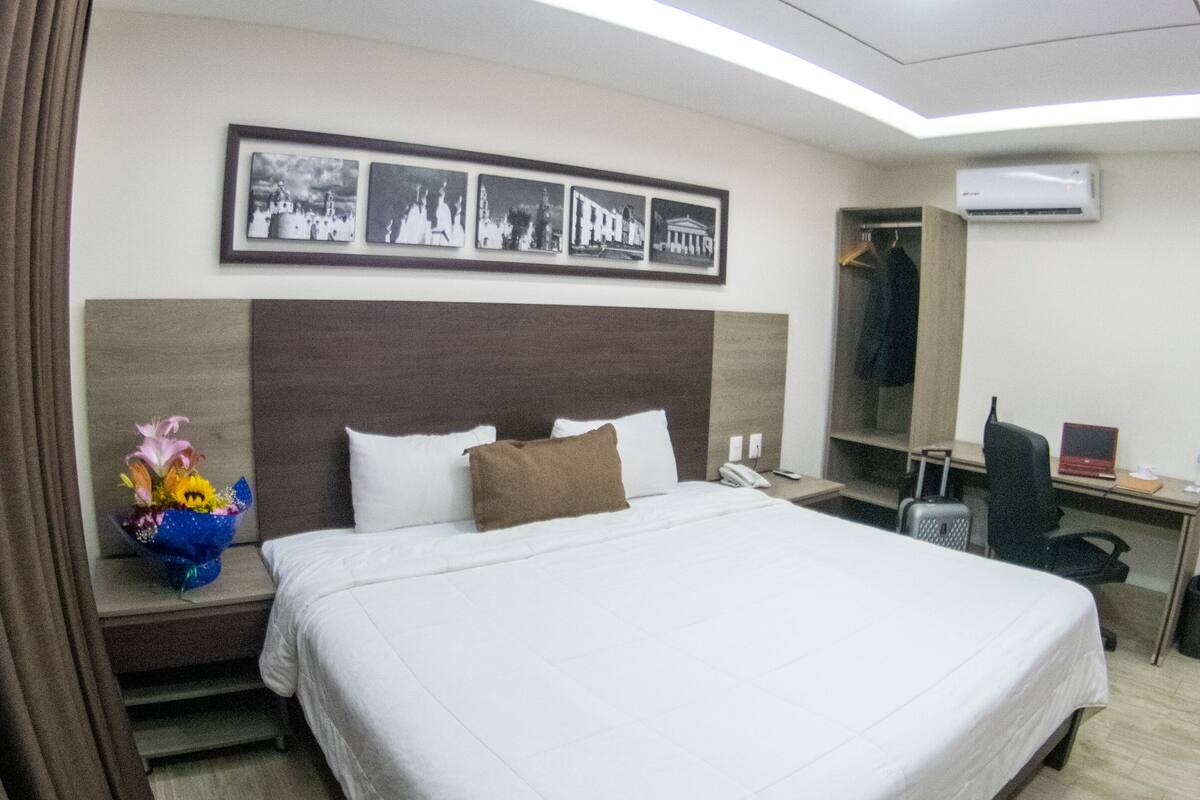 A hotel room with a large bed, a desk with a chair, a wardrobe, and a wall-mounted air conditioner.