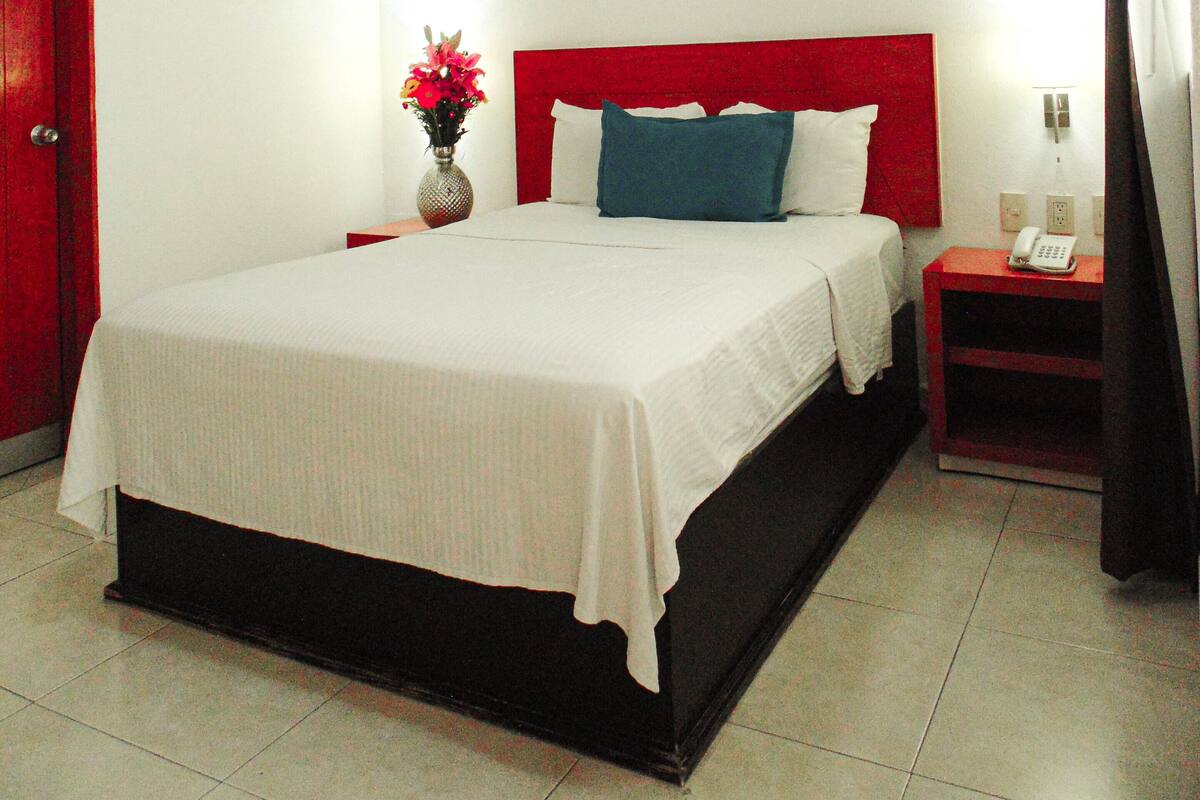 A hotel room with a bed, a bedside table with a phone, a red headboard, a ceiling fan, and a framed picture on the wall.