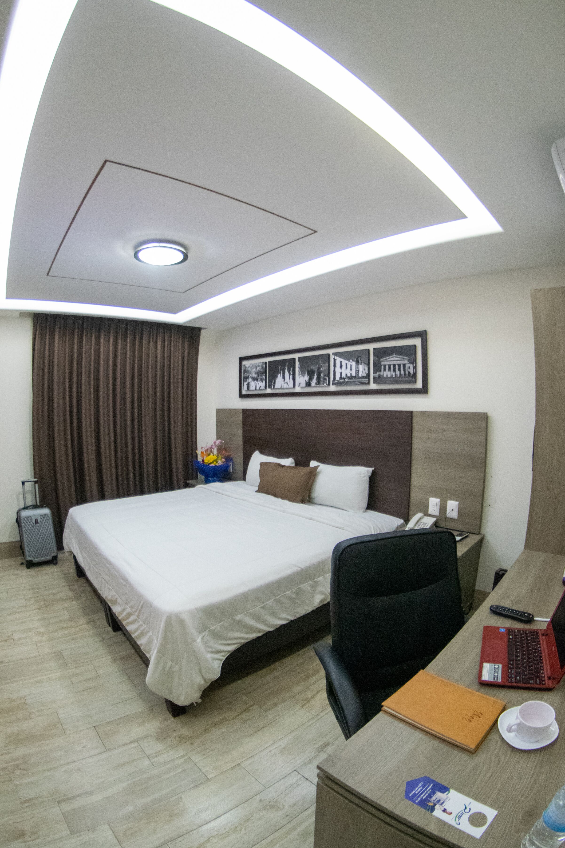 Habitación de hotel con una cama grande, un escritorio con una computadora portátil, una silla, una maleta y un cuadro en la pared.