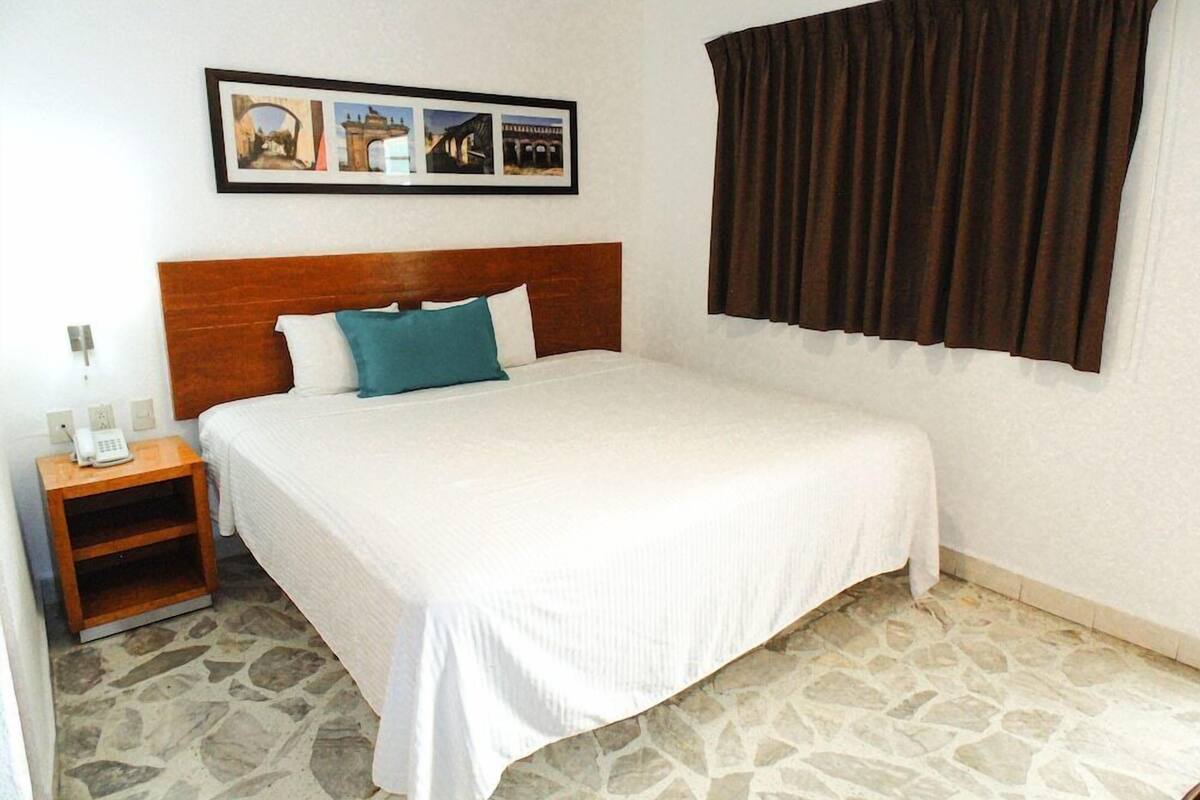 A bedroom with a bed, a nightstand, a telephone, a framed picture on the wall, and a window with curtains.