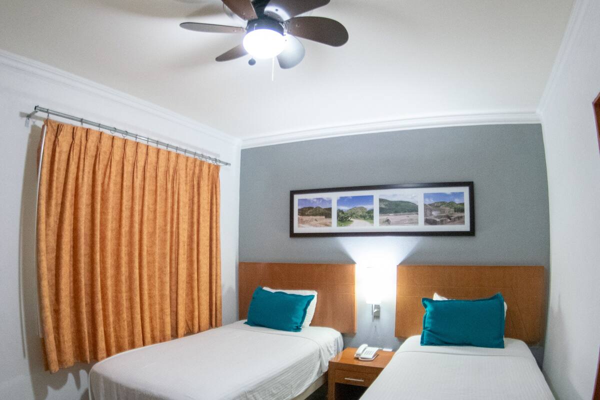 A hotel room with two single beds, a ceiling fan, a nightstand with a lamp, and a framed picture on the wall.