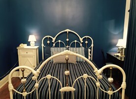 Una cama con estructura metálica blanca y ropa de cama a rayas, una mesita de noche con lámpara y otro apoyo para objetos con una lámpara.