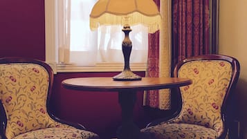 A room with two patterned armchairs, a round table with a lamp, and a window with sheer curtains.