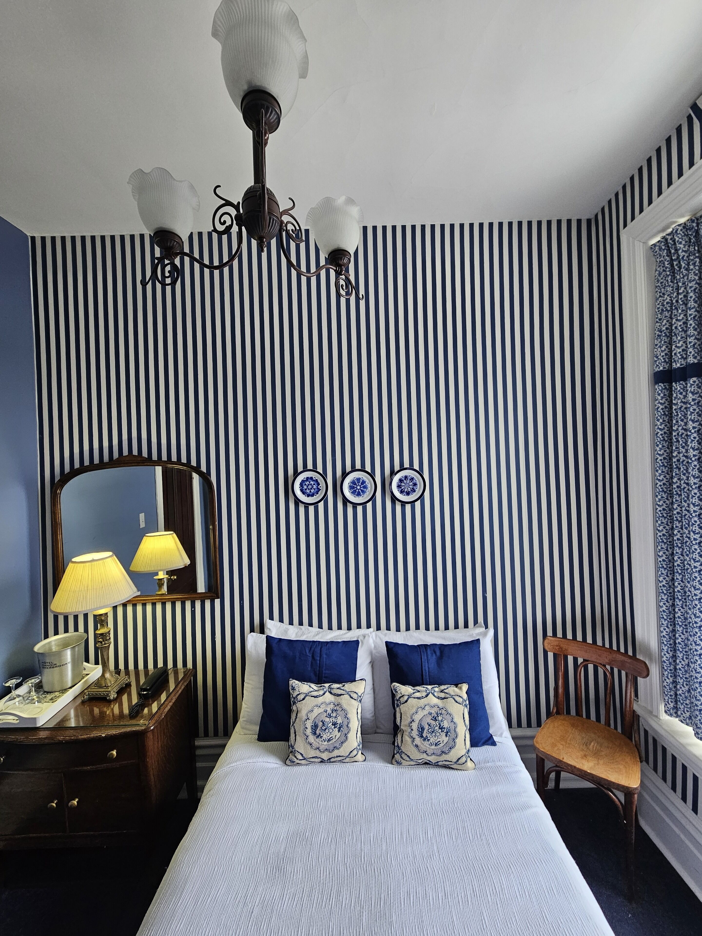 A bedroom with a striped wall, a bed with blue and white bedding, a bedside table with a lamp, a mirror, and a window with curtains.