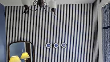 A bedroom with a striped wall, a bed with blue and white bedding, a bedside table with a lamp, a mirror, and a window with curtains.