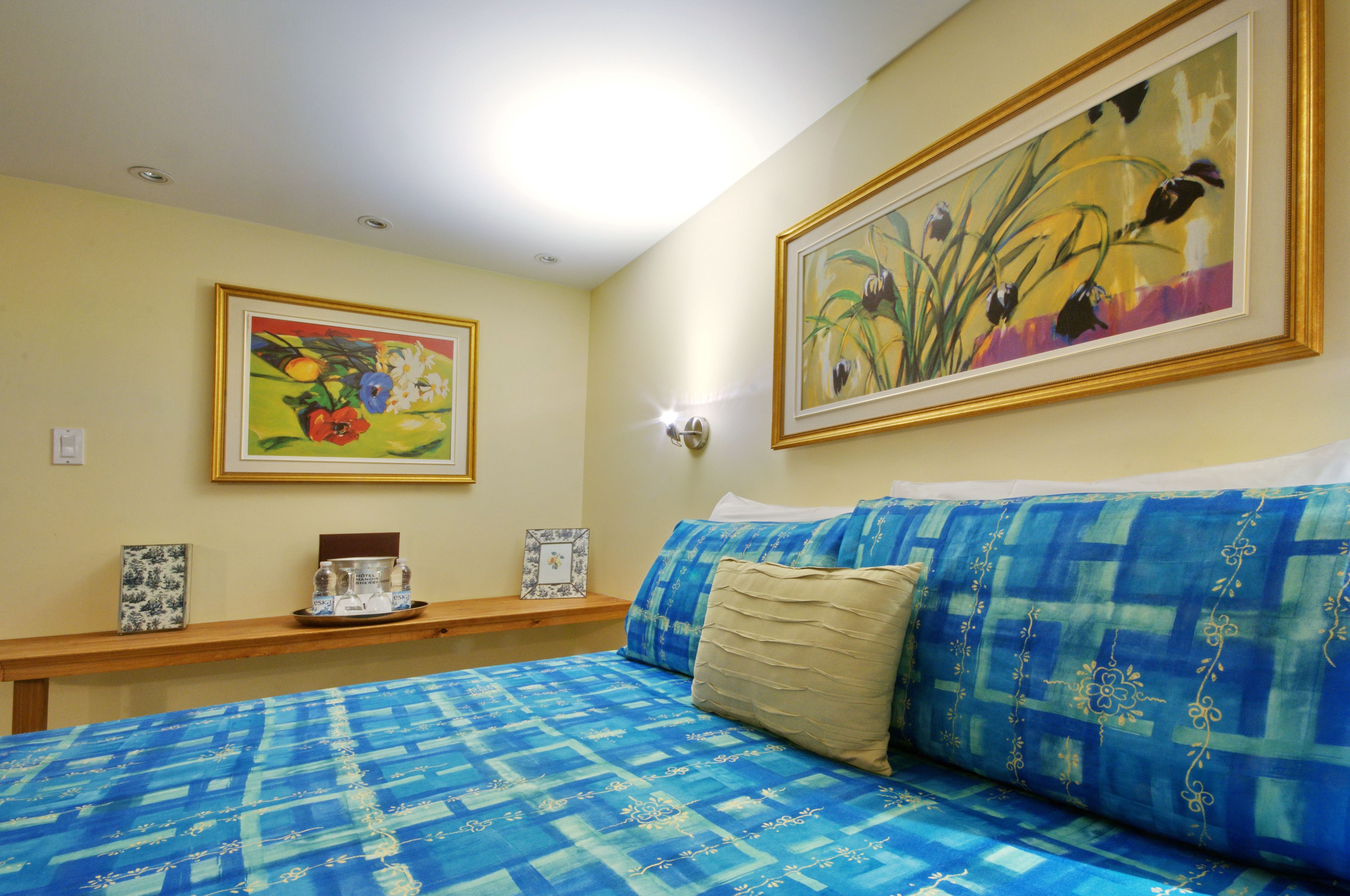 A bedroom with a blue patterned bedspread, a beige pillow, and two framed floral paintings on the wall.