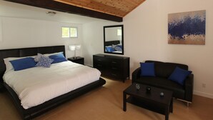 A bedroom with a large bed, a black dresser, a black sofa, a coffee table, a mirror, and a painting on the wall. - Baywatch Hotel & Marina (Hampton Bays)