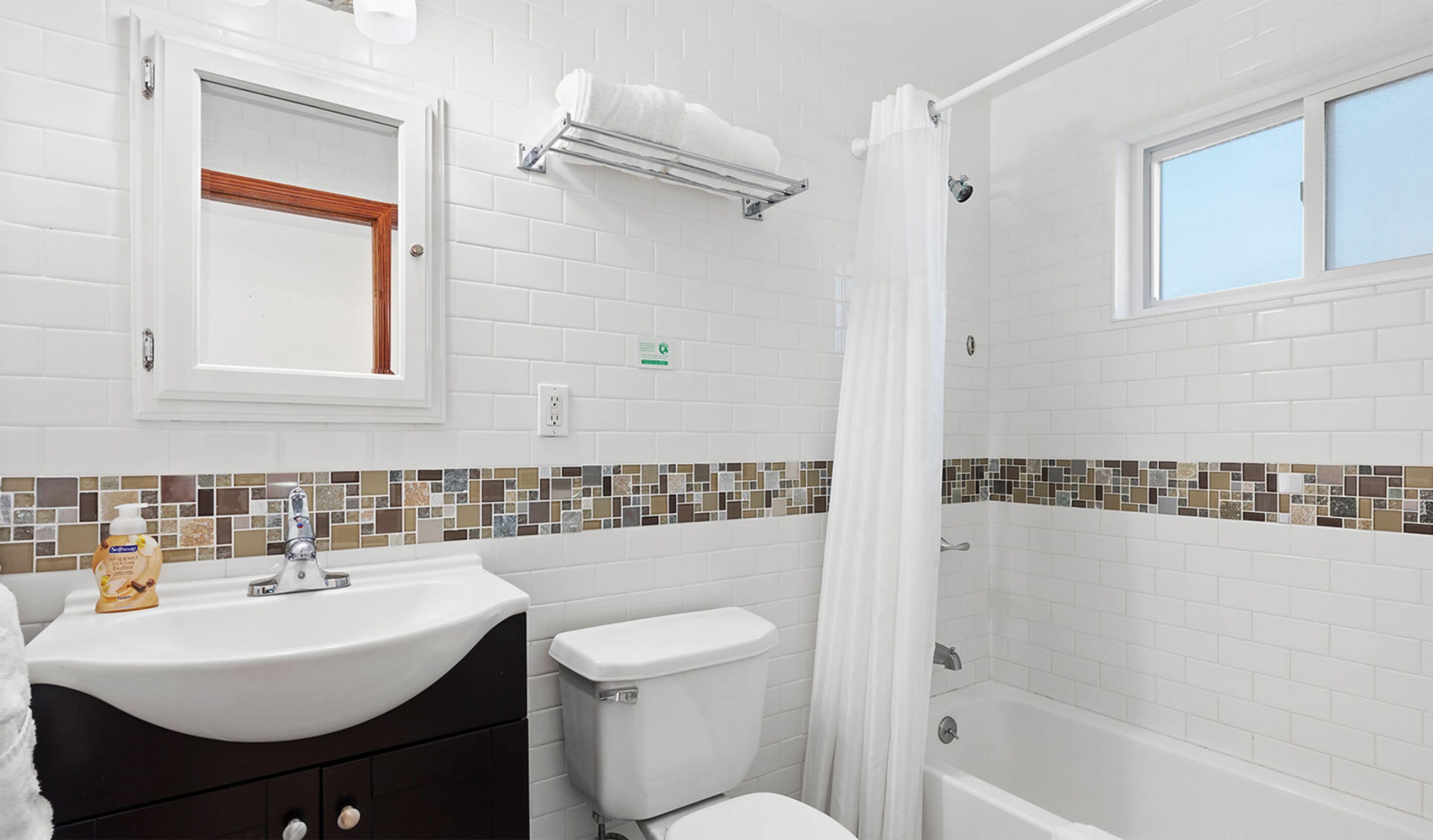 Un baño con lavamanos, inodoro y bañera blancos. Hay una ventana sobre la bañera y un espejo sobre el lavamanos.