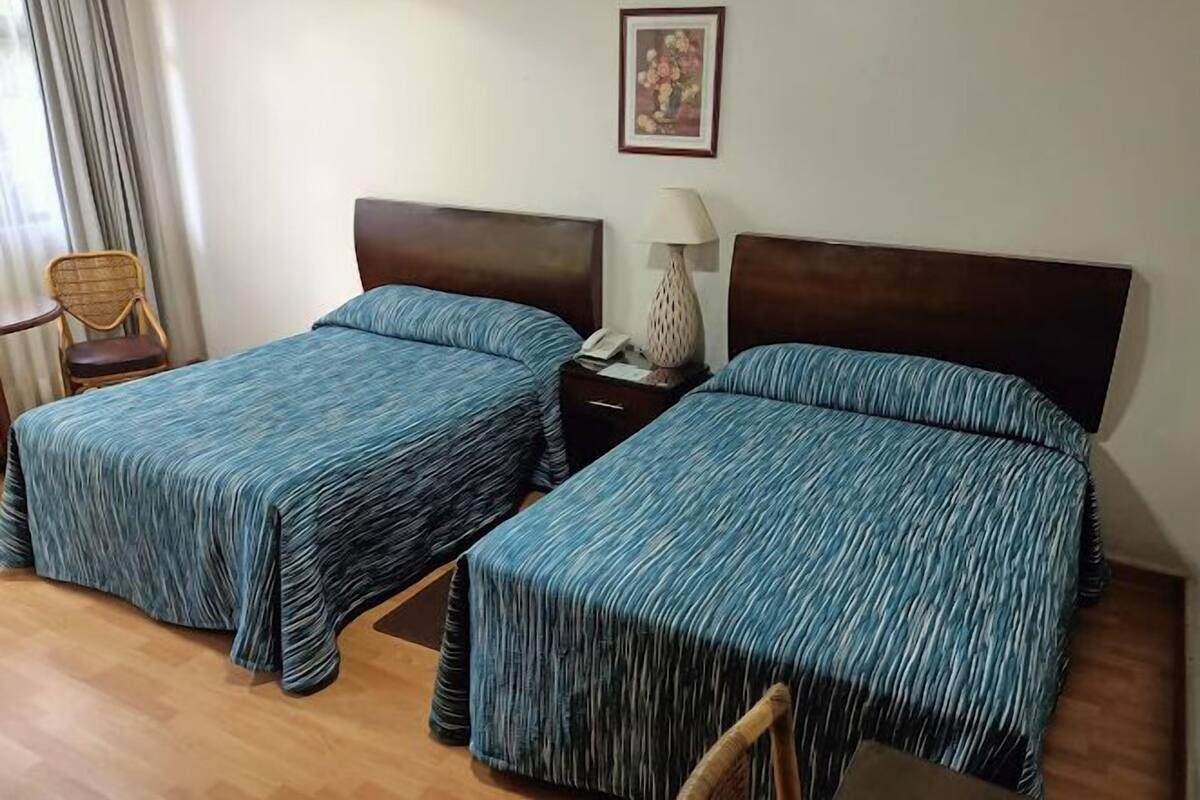 Hotellihuone, jossa on kaksi sänkyä, tuoli, yöpöytä lampun kanssa ja seinällä kehystetty kuva.