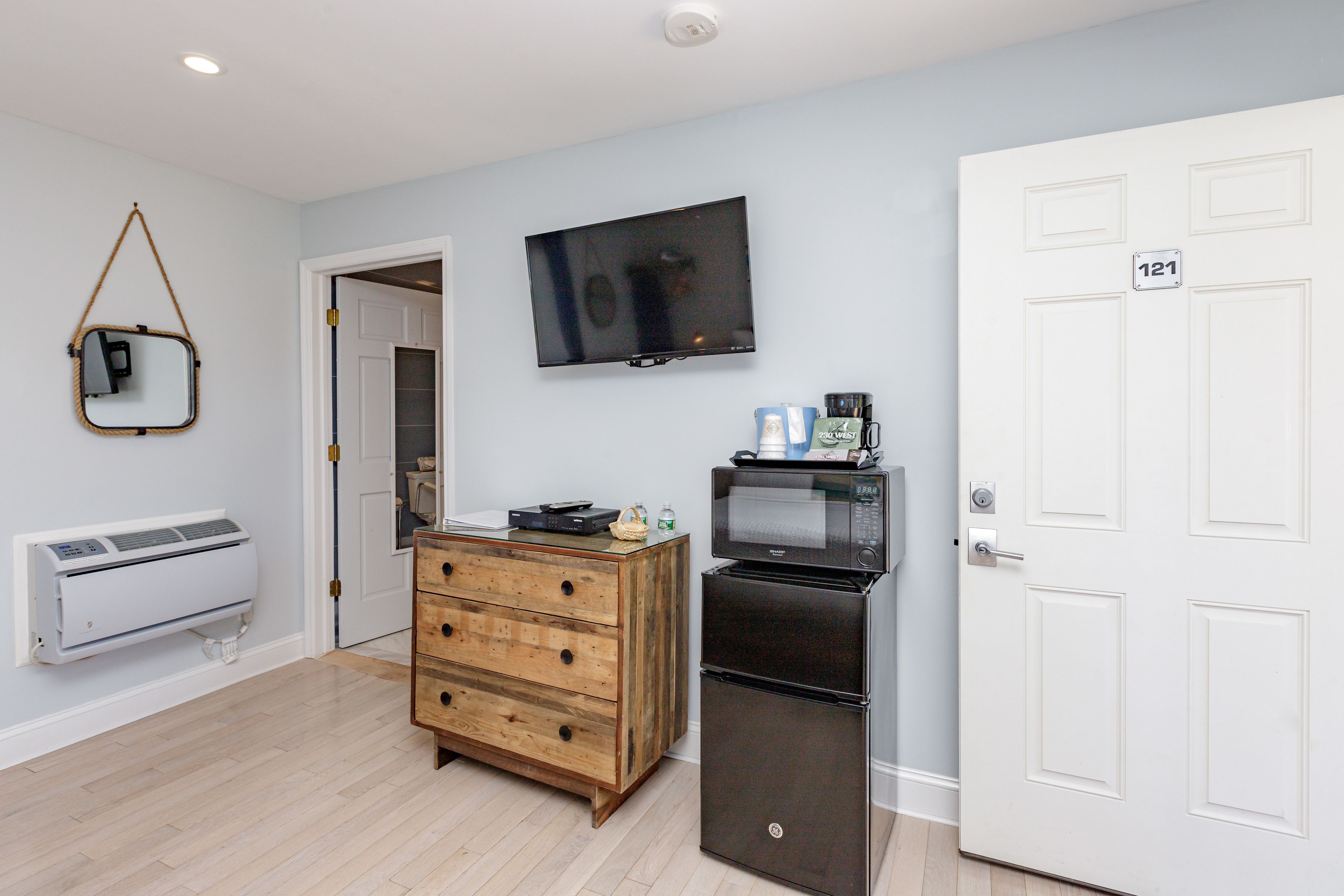 A room with a wooden dresser, a black refrigerator, a microwave, a television, and a white door with a number plate.