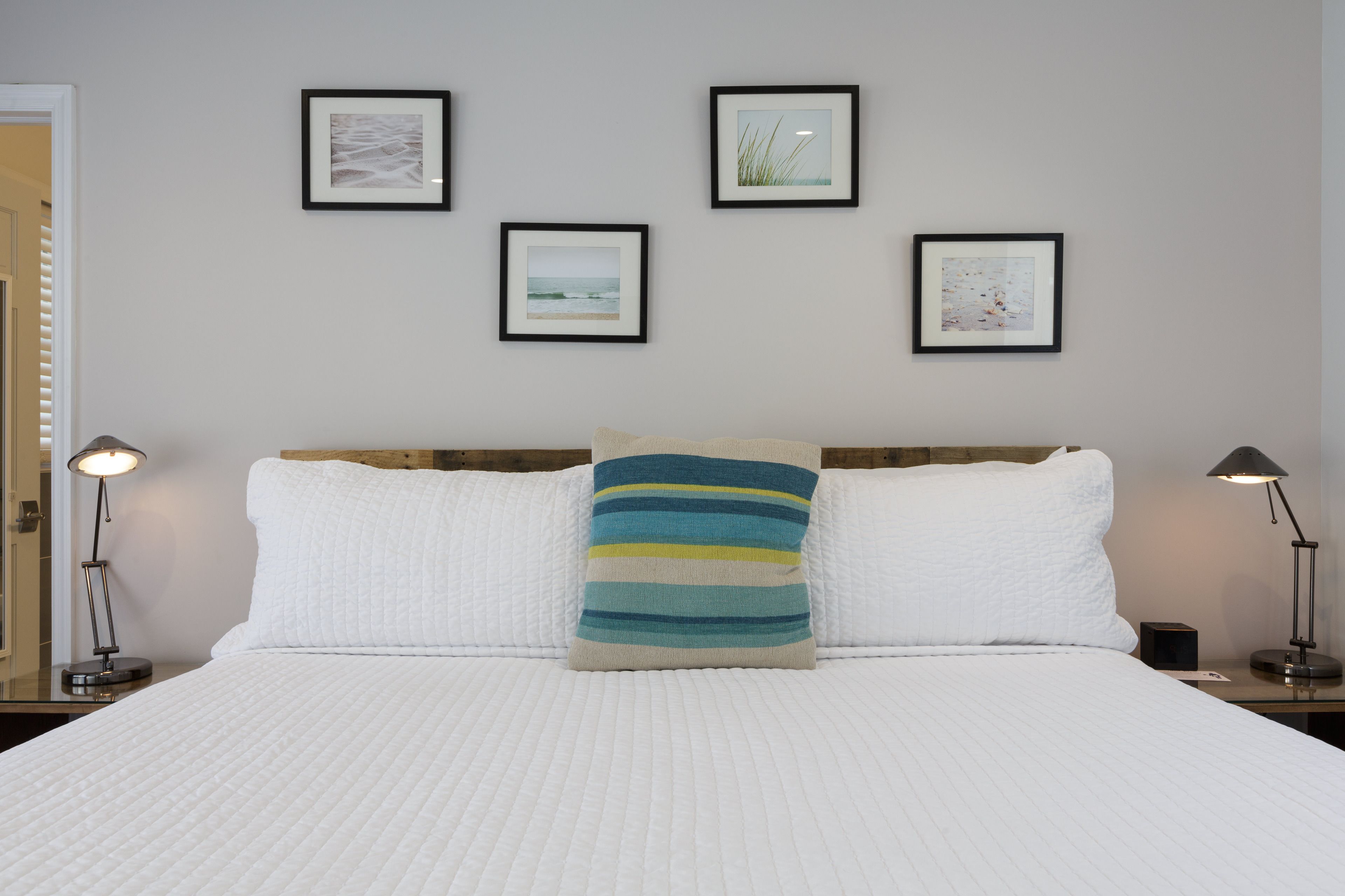 A neatly made bed with white bedding and a striped pillow, flanked by two bedside lamps. Four framed pictures adorn the wall above the bed.