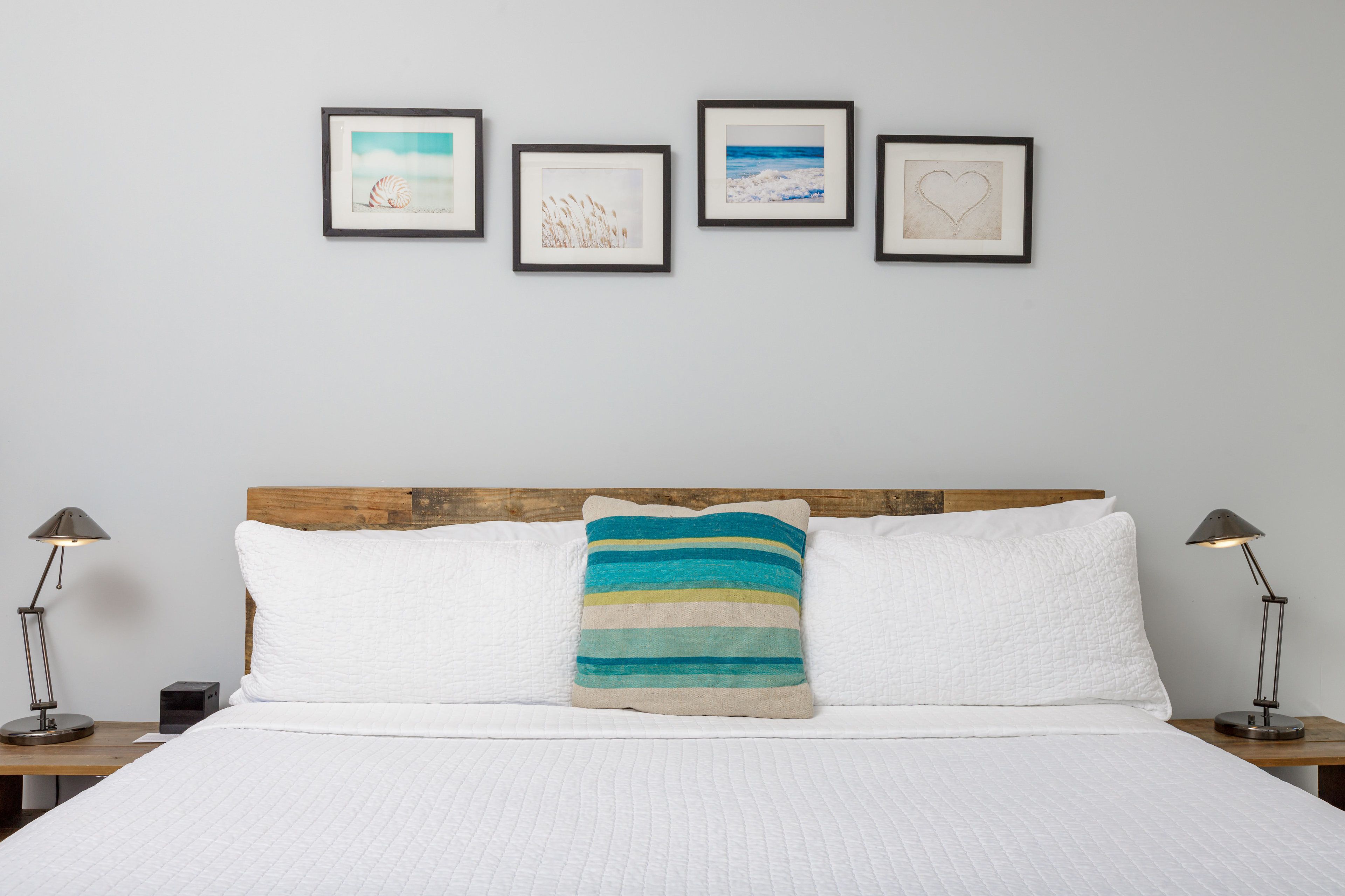 Ein ordentlich bezogenes Bett mit weißer Bettwäsche, einem Holz-Kopfteil und einem farbenfrohen, gestreiften Kopfkissen. Vier gerahmte Kunstwerke hängen an der Wand über dem Bett.
