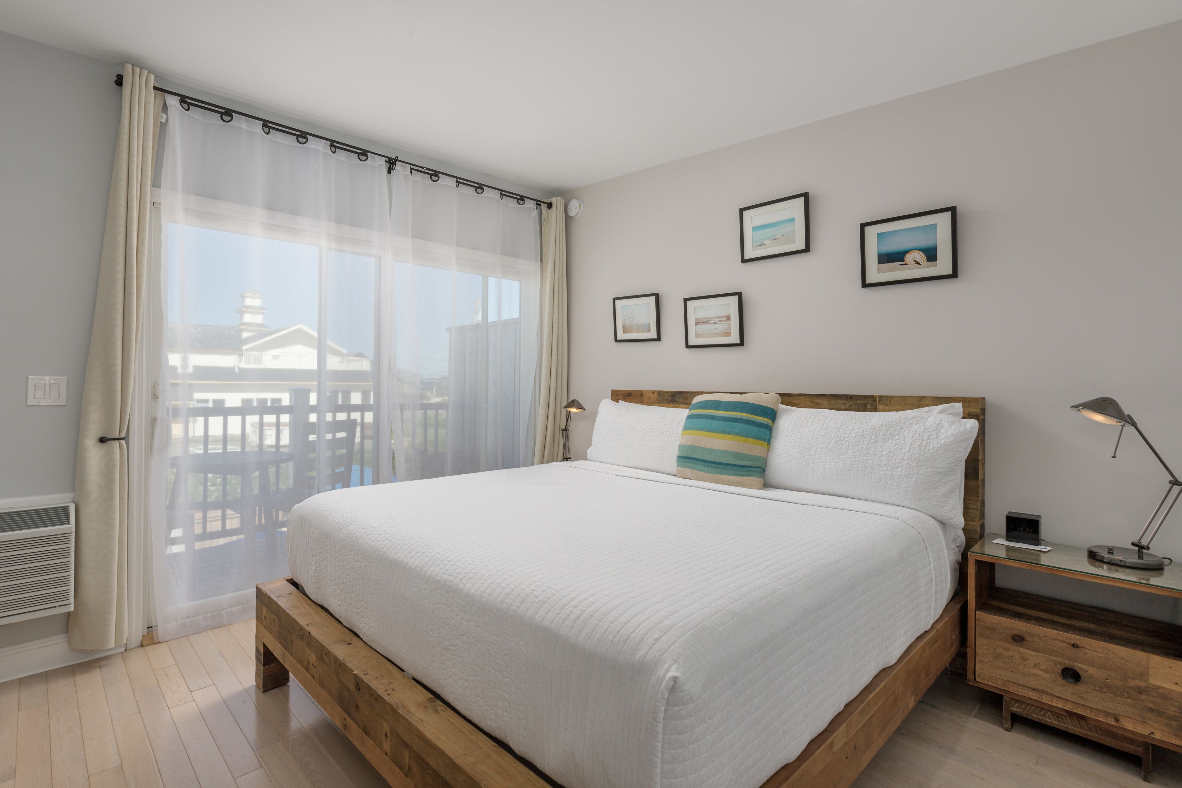 Un dormitorio con una cama grande, cabecera de madera, mesita de noche con lámpara y vistas a un balcón con barandilla.