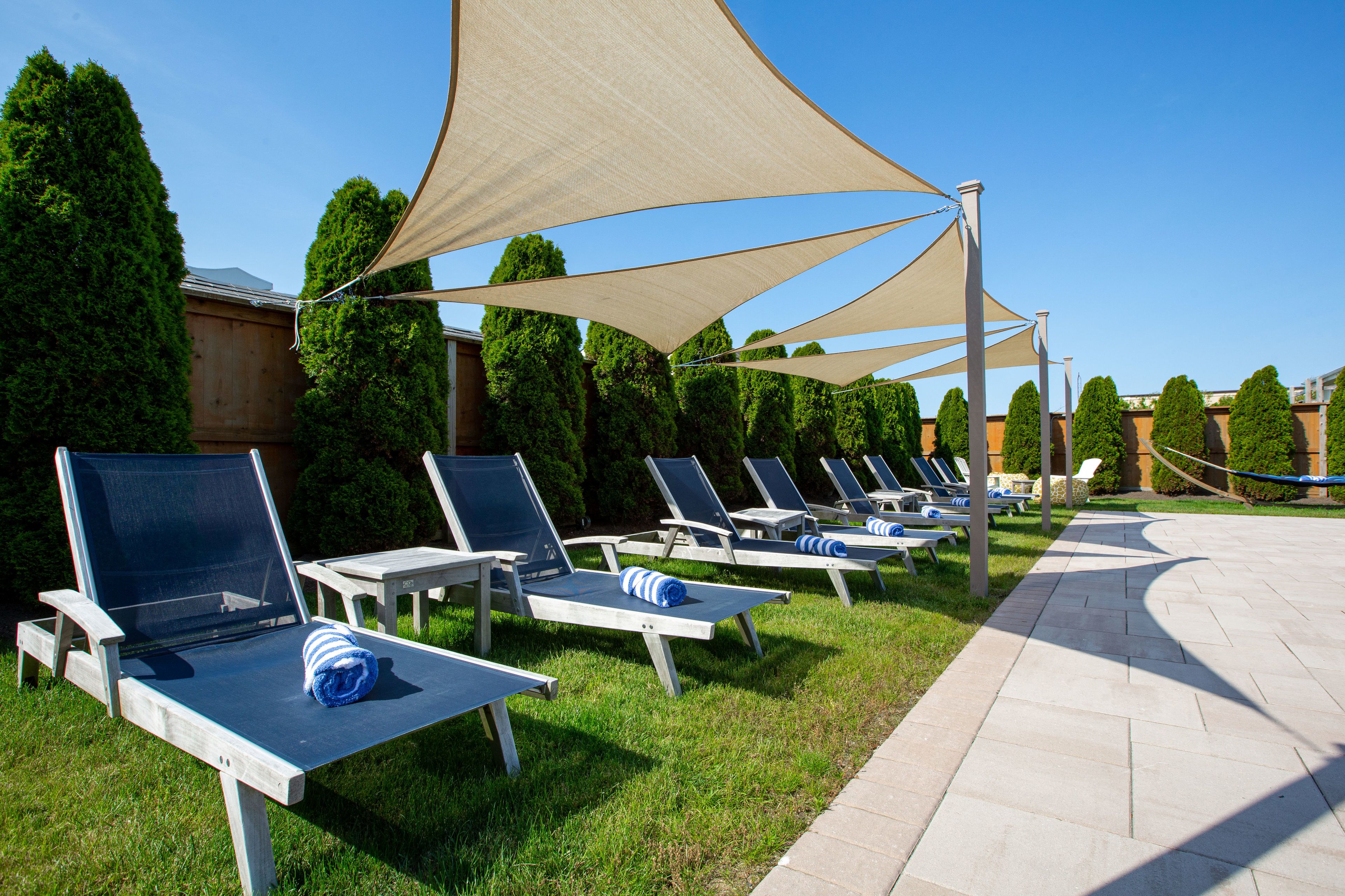Une rangée de chaises longues surmontées de parasols beiges, un chemin pavé et des haies soigneusement taillées.