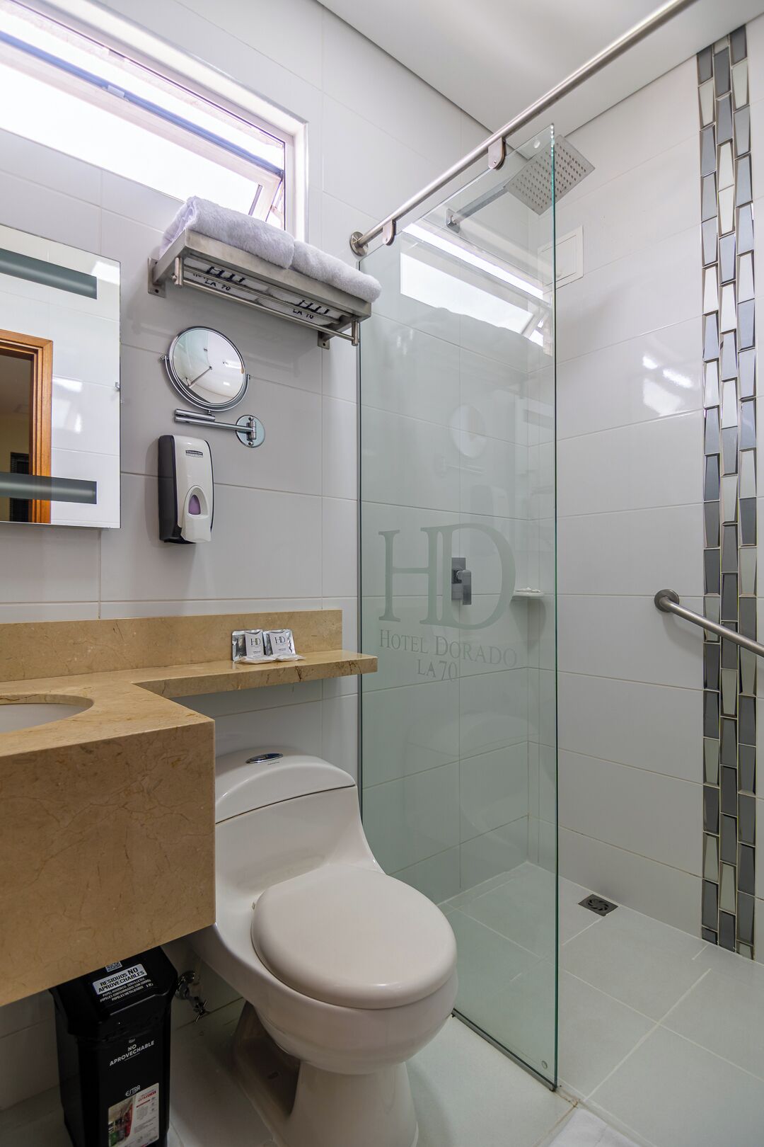 Et moderne badeværelse med glasvæg omkring bruseren, toilet, vask med sæbedispenser, et ur og et spejl.