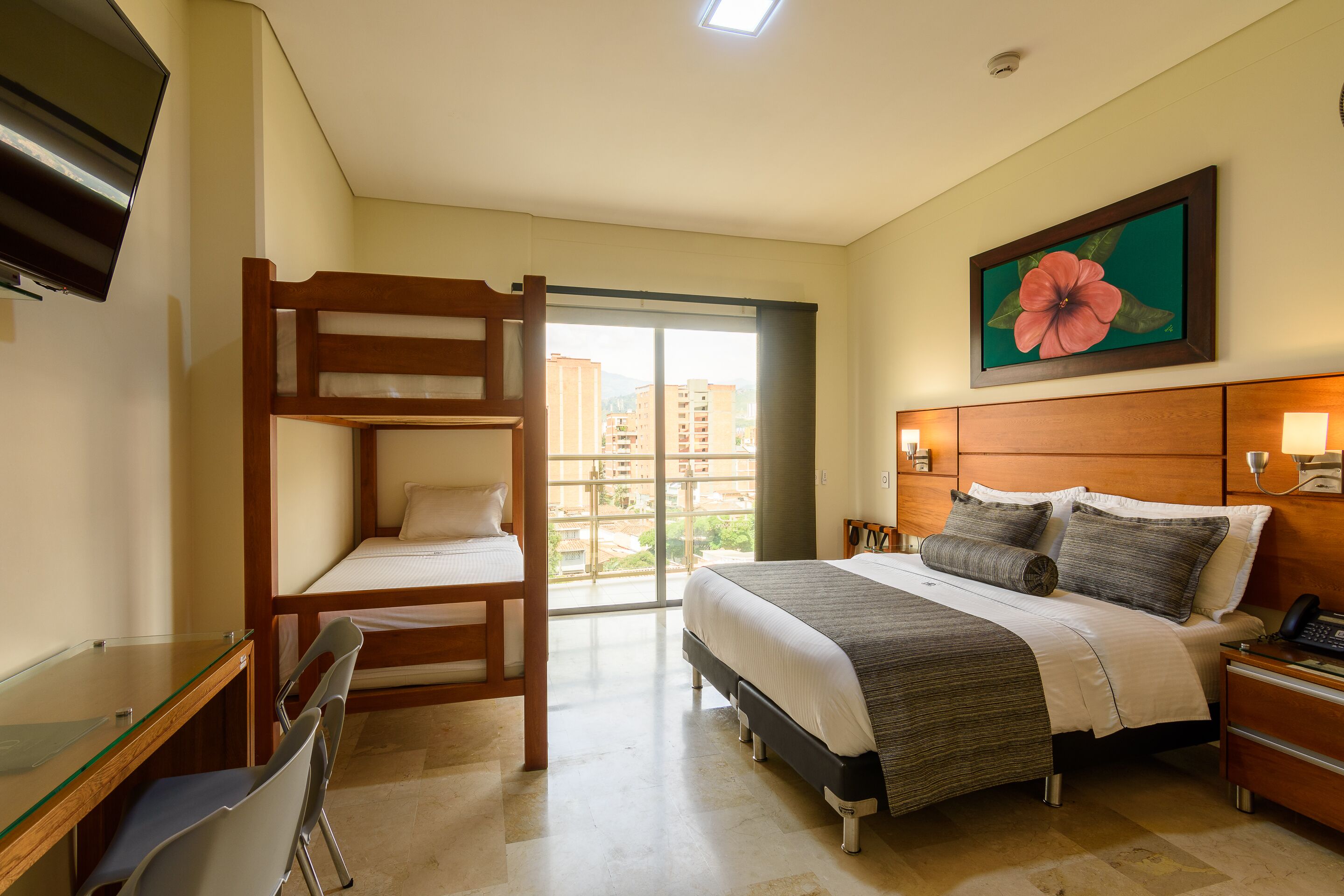 A hotel room with a bunk bed, a desk, a chair, a bed with pillows, a nightstand, a lamp, a painting, and a view of buildings through the window.