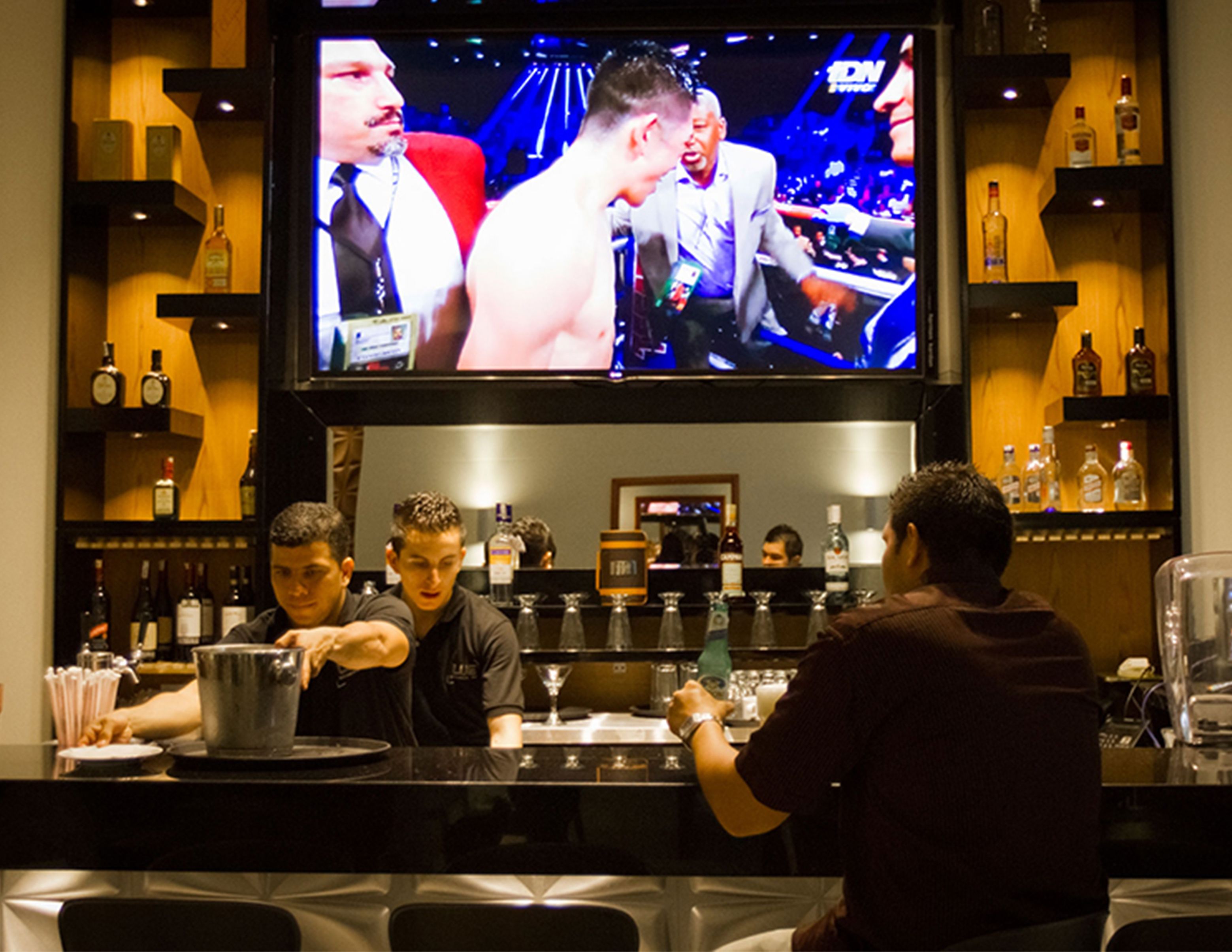 A bar area with a television displaying a boxing match, shelves with bottles, and a bartender preparing drinks.