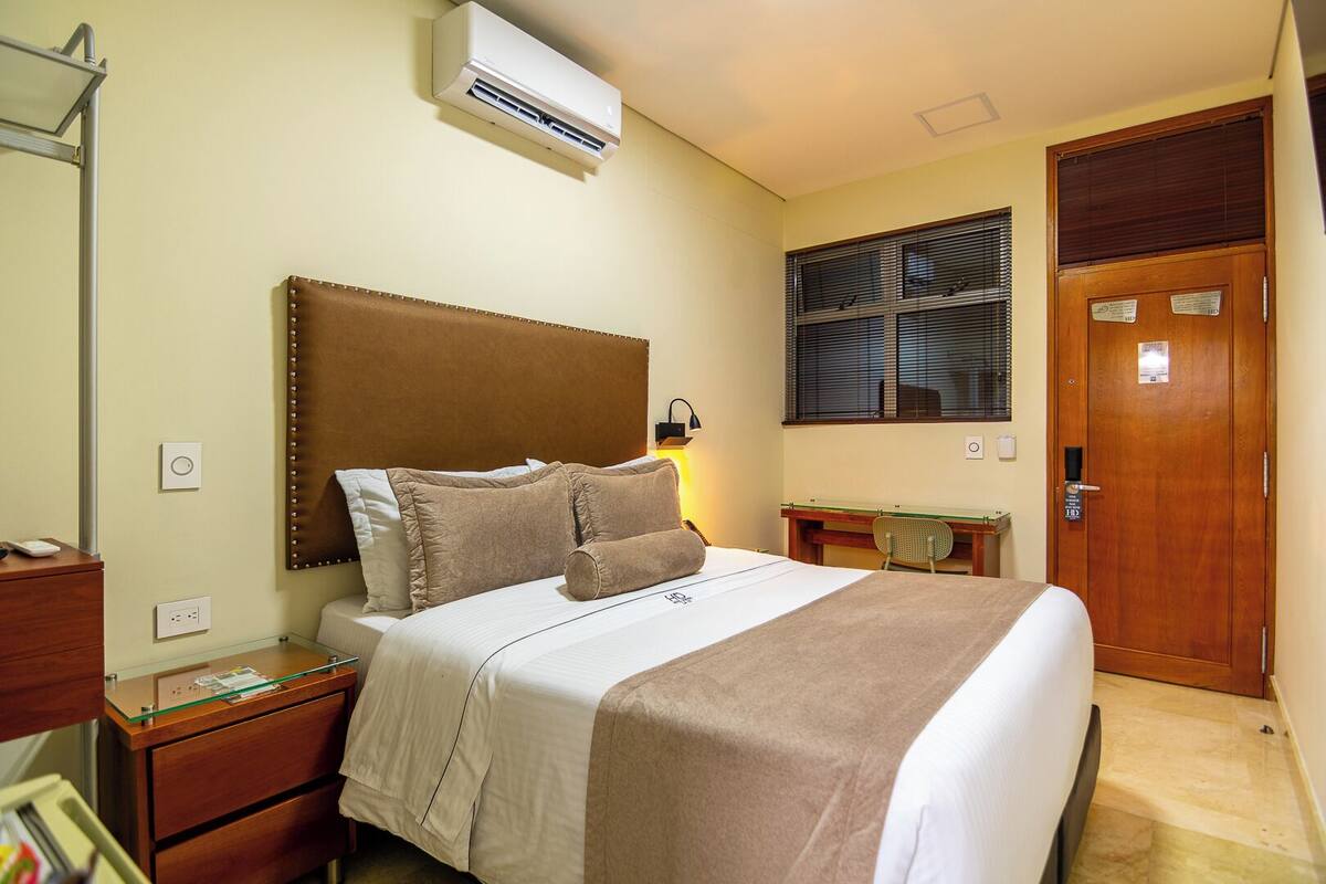 Een hotelkamer met een bed, nachtkastje, lamp, airconditioning, raam en een deur.