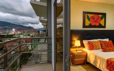 A hotel room with a balcony overlooking a cityscape, a bed with patterned pillows, and a painting of a flower on the wall.