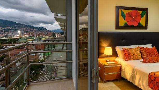 A hotel room with a balcony overlooking a cityscape, a bed with patterned pillows, and a painting of a flower on the wall.