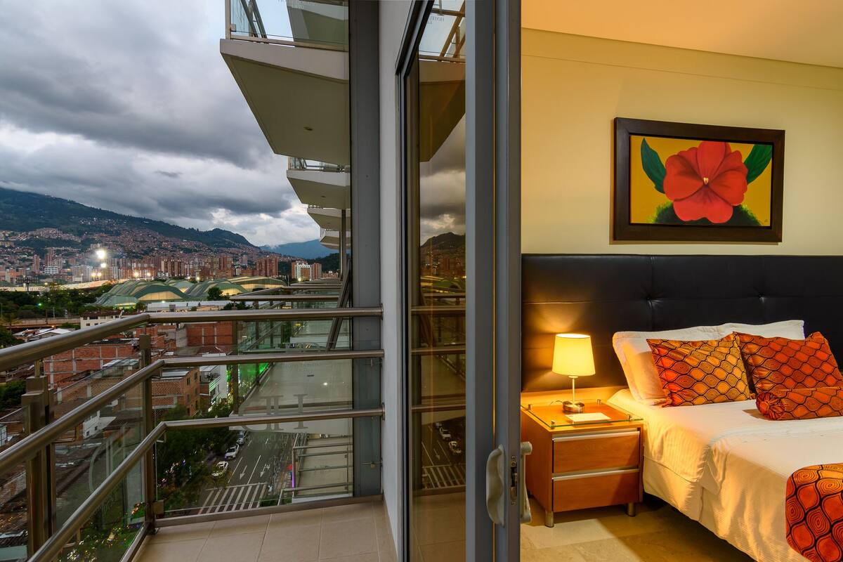 Hotelkamer met balkon en uitzicht over de stad, een bed met geborduurde kussens en een bloemschilderij aan de muur.