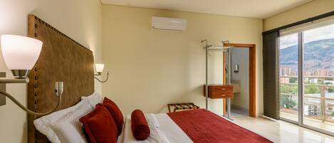 Habitación de hotel con una cama grande, una lámpara de noche, un equipo de aire acondicionado, un baño con puerta de vidrio y vistas a la ciudad.