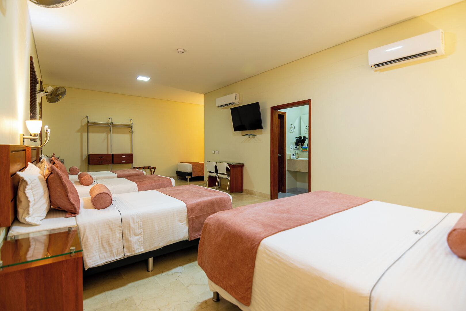 Habitación de hotel con tres camas, un escritorio con silla, un televisor y un aire acondicionado.
