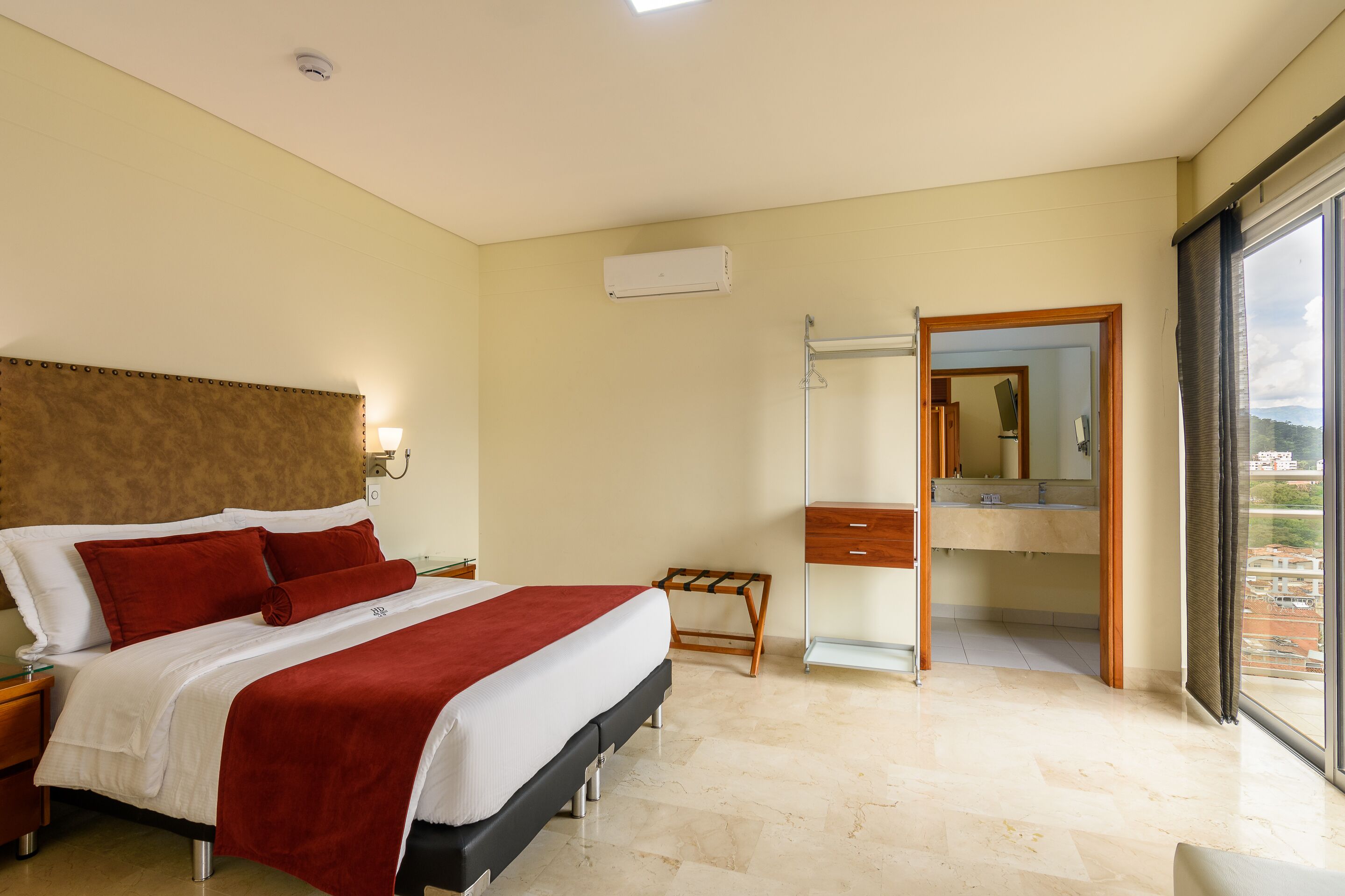 Habitación de hotel con cama, mesita de noche, espejo, baño con lavamanos y vista a la ciudad.