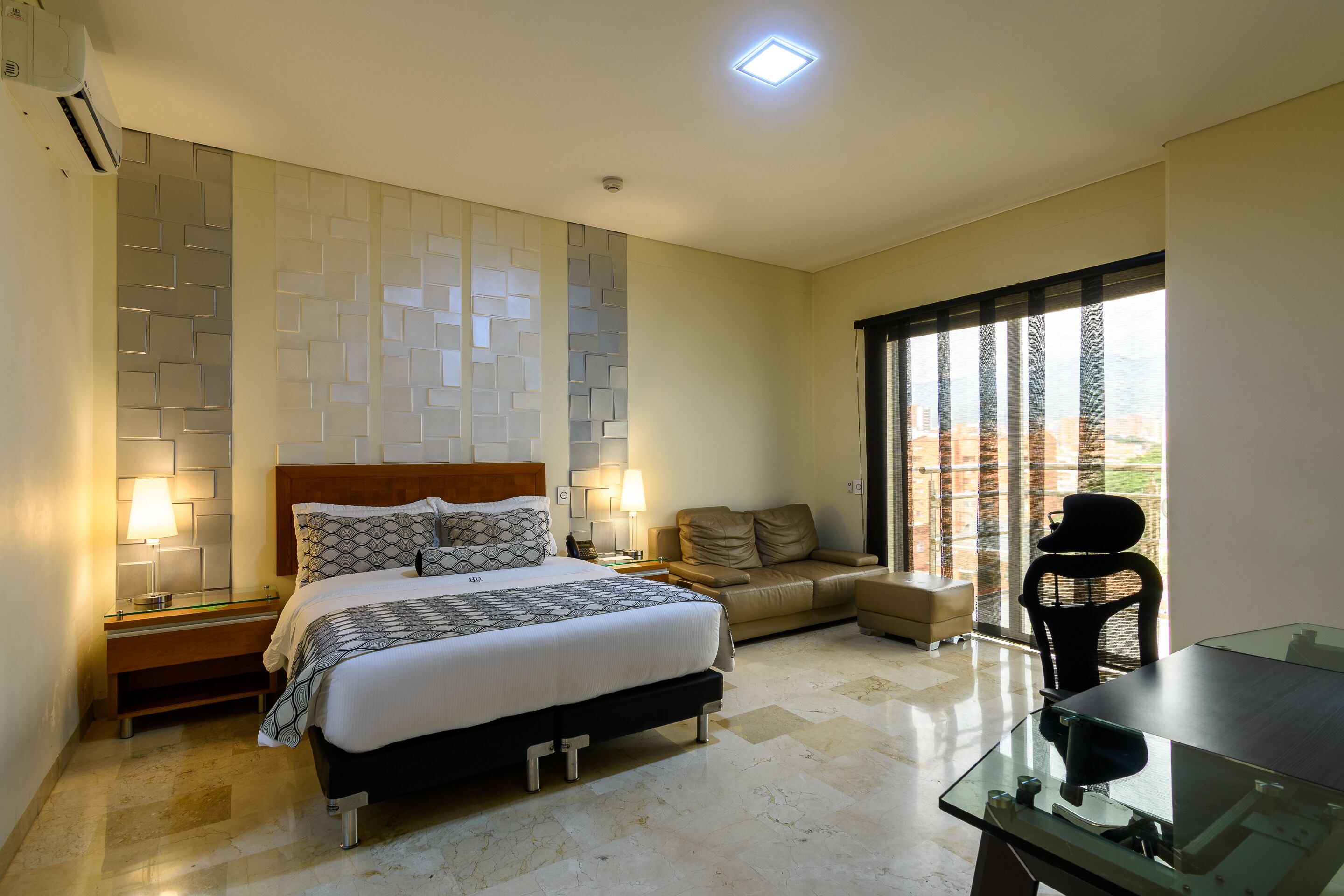 Una habitación de hotel moderna con una cama grande, un sofá, una mesa de comedor y vistas a la ciudad a través de amplias ventanas.