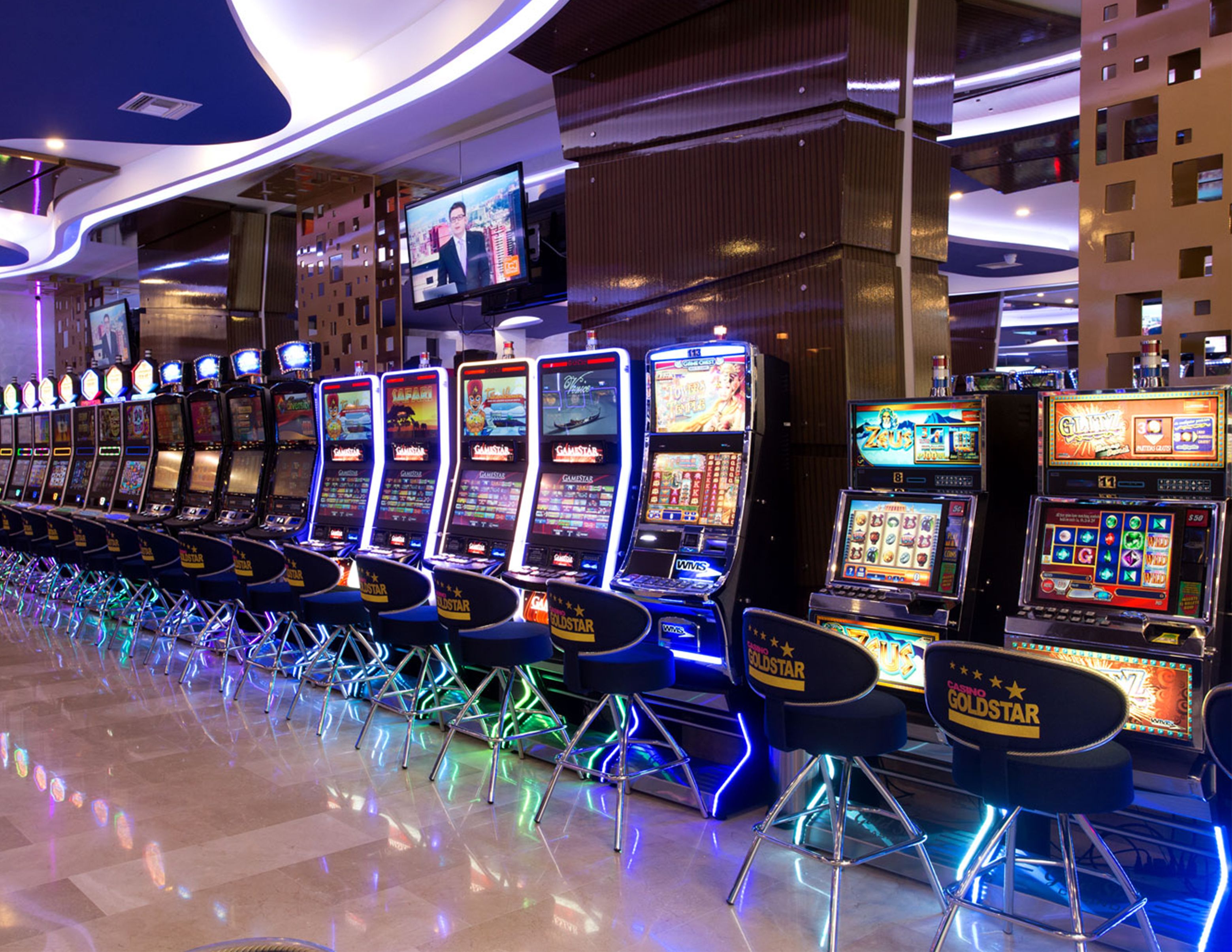 A row of slot machines with neon lights and a television screen above them.