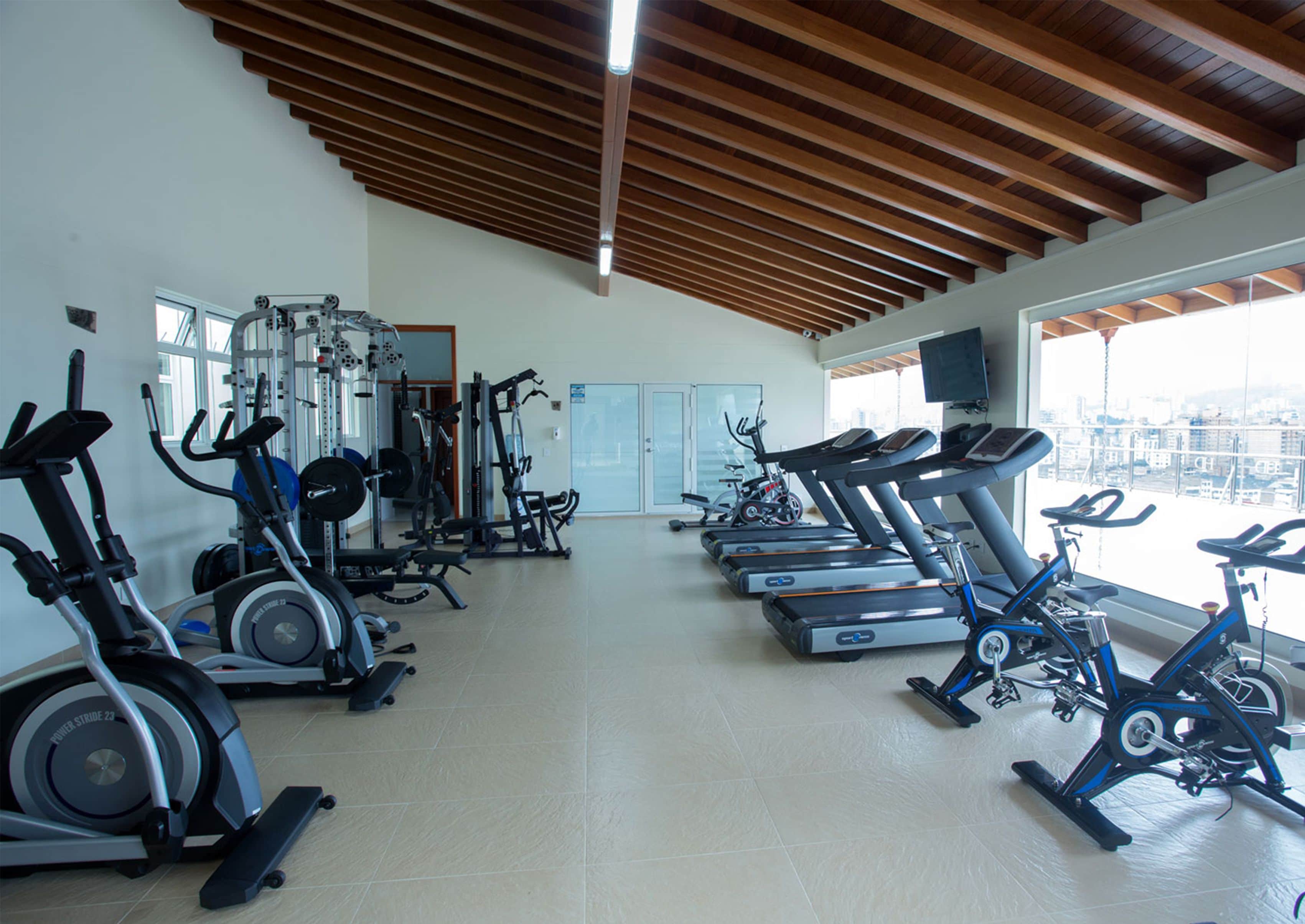 A gym with various exercise equipment, a large window with a city view, and a wooden ceiling.