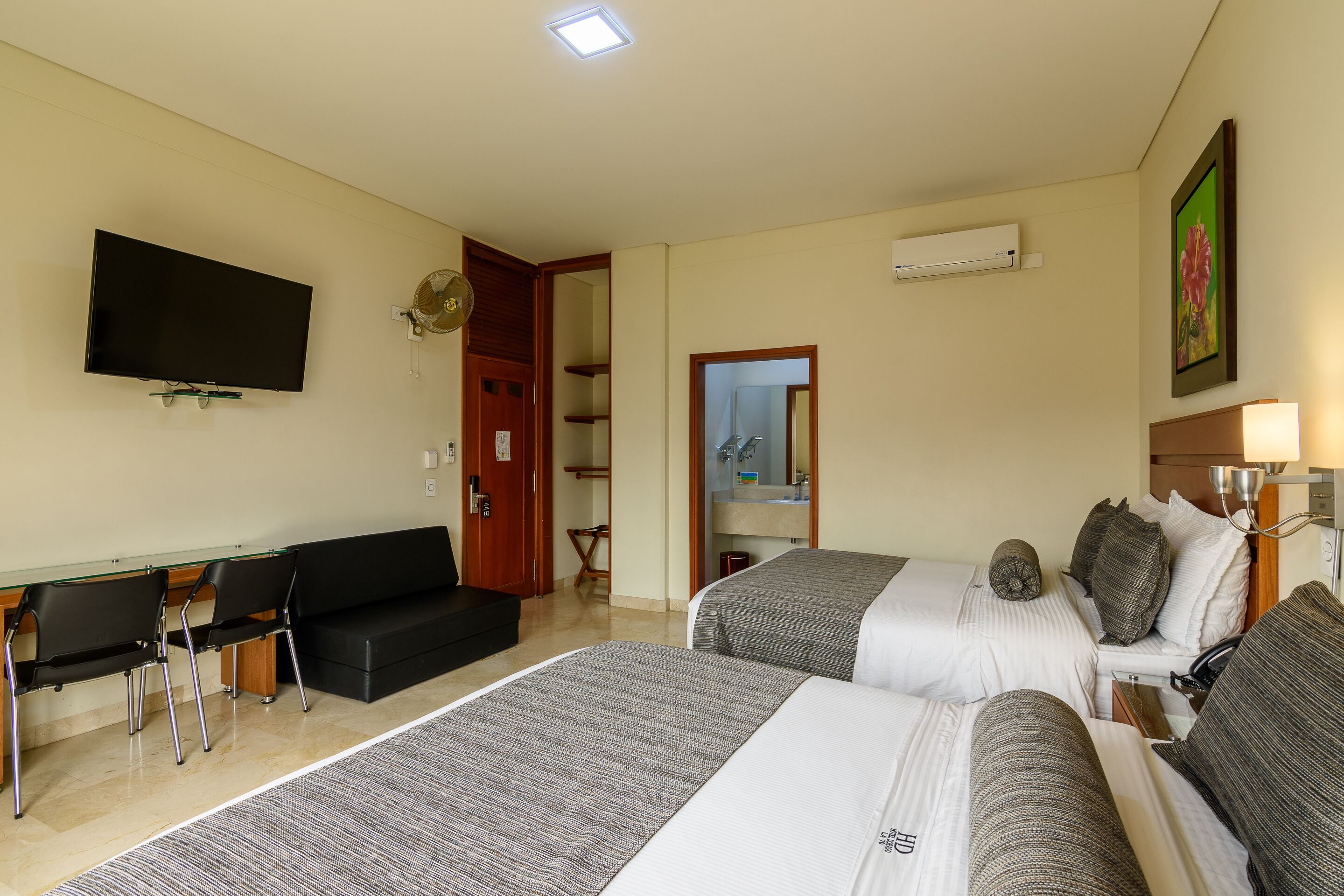 A hotel room with two beds, a desk with chairs, a TV, and a bathroom visible through an open door.