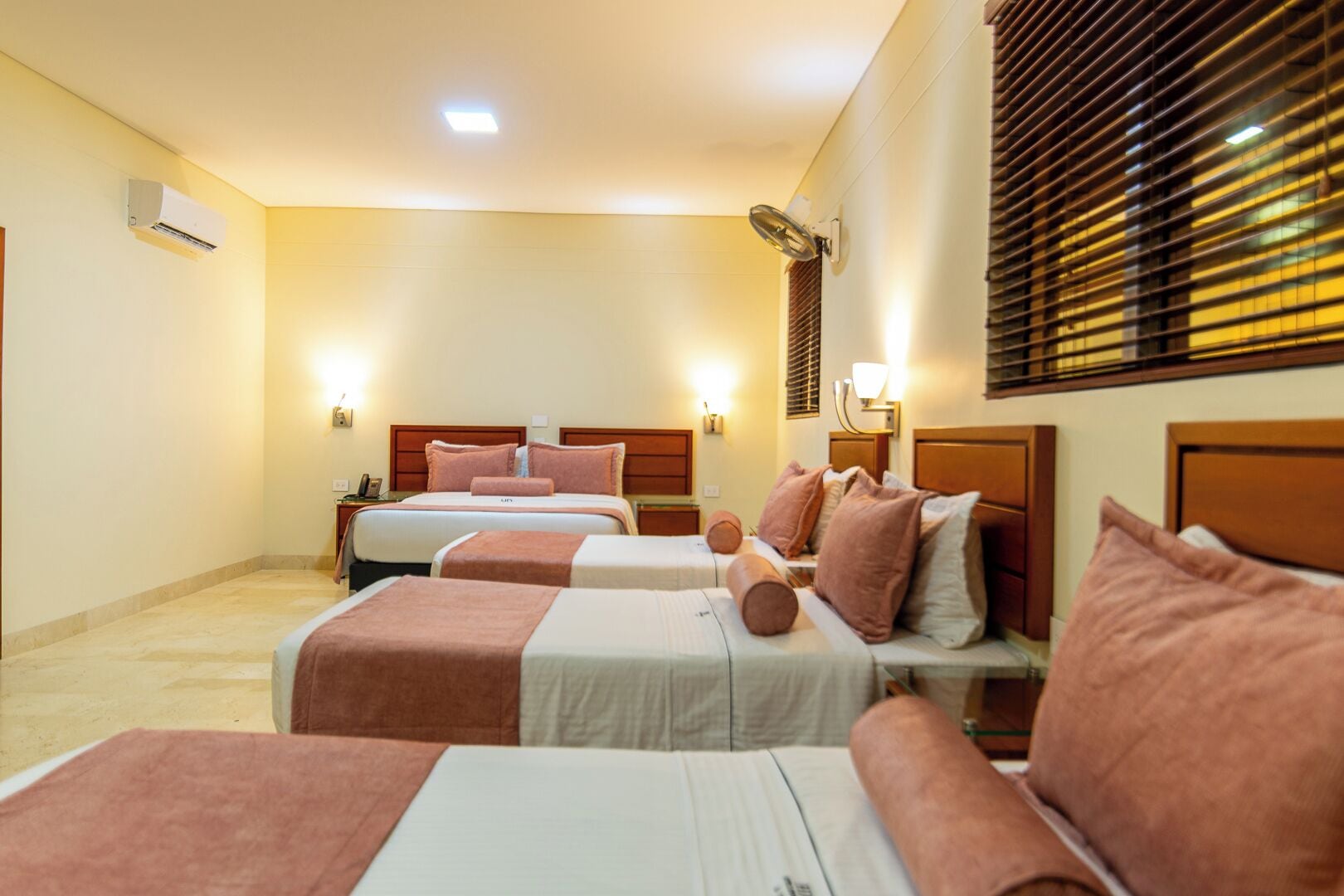 A hotel room with three beds, a wall-mounted air conditioner, and bedside lamps.