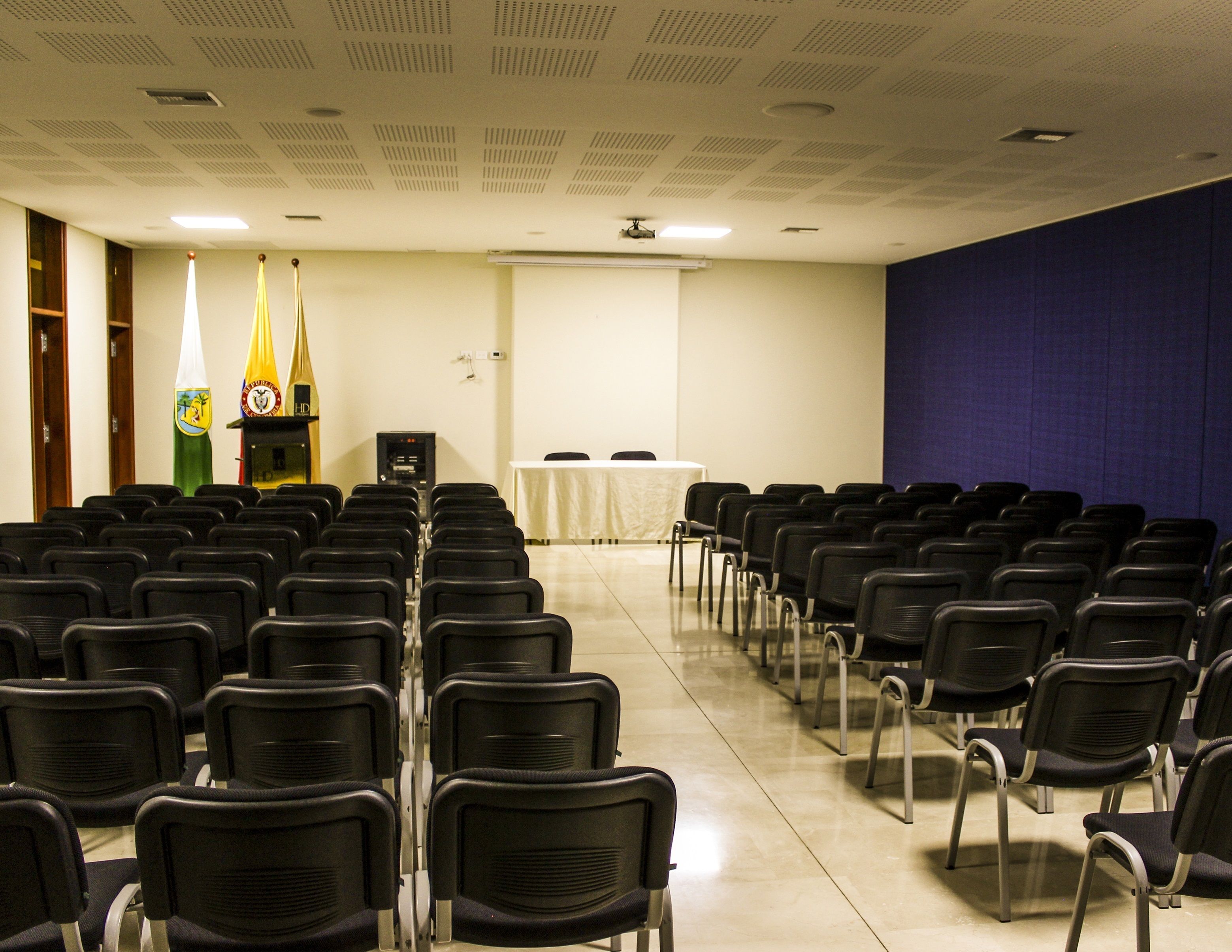 Une salle de conférence avec des rangées de chaises noires, une table blanche et deux drapeaux sur le mur de gauche.