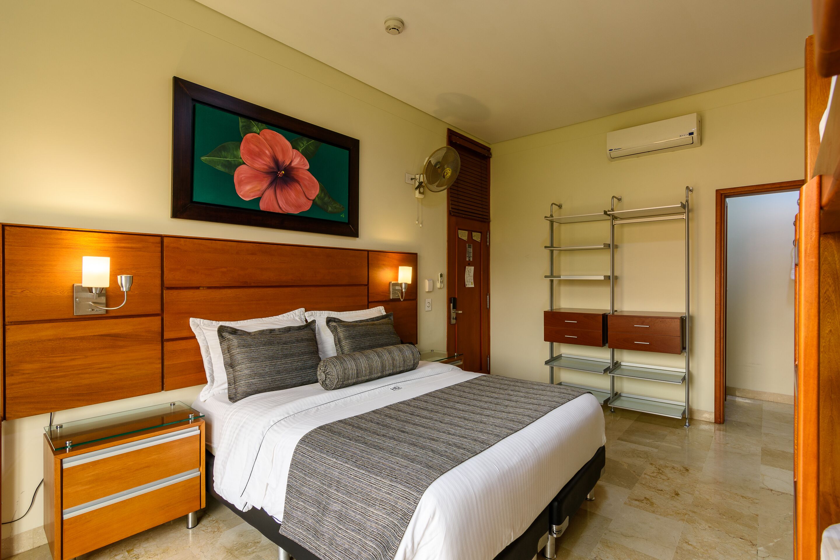 A hotel room with a large bed, wooden headboard, bedside tables, a mirror, a wardrobe, and a large floral painting on the wall.