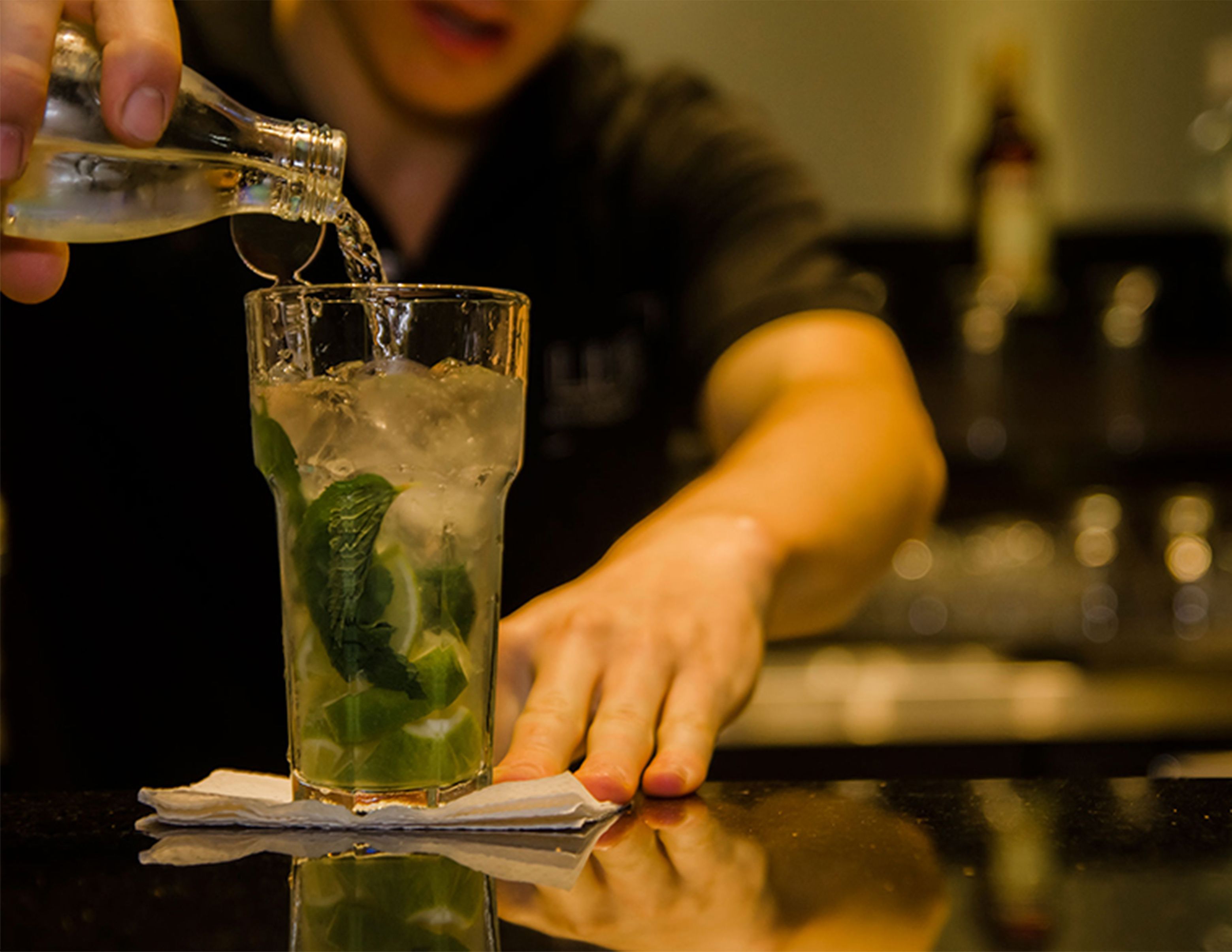 A person pouring a clear liquid into a glass with ice and mint leaves.