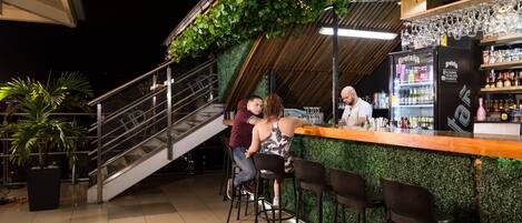 Zona de bar al aire libre con una escalera, taburetes y un cantinero.